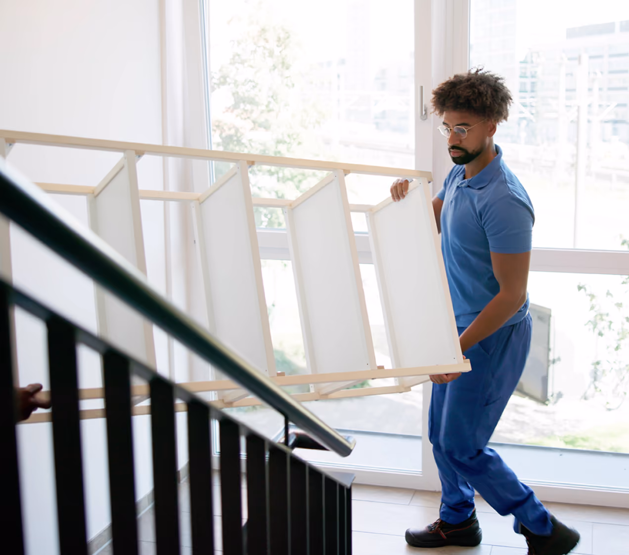 Man in blue shirt carrying a flat-pack furniture frame up a stairwell near a large window.