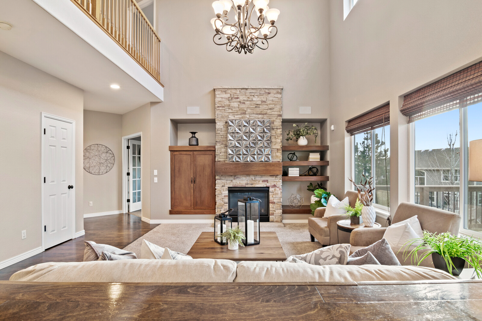 Bright living room with stone fireplace, wooden shelves, beige sofa, and large windows with brown blinds.