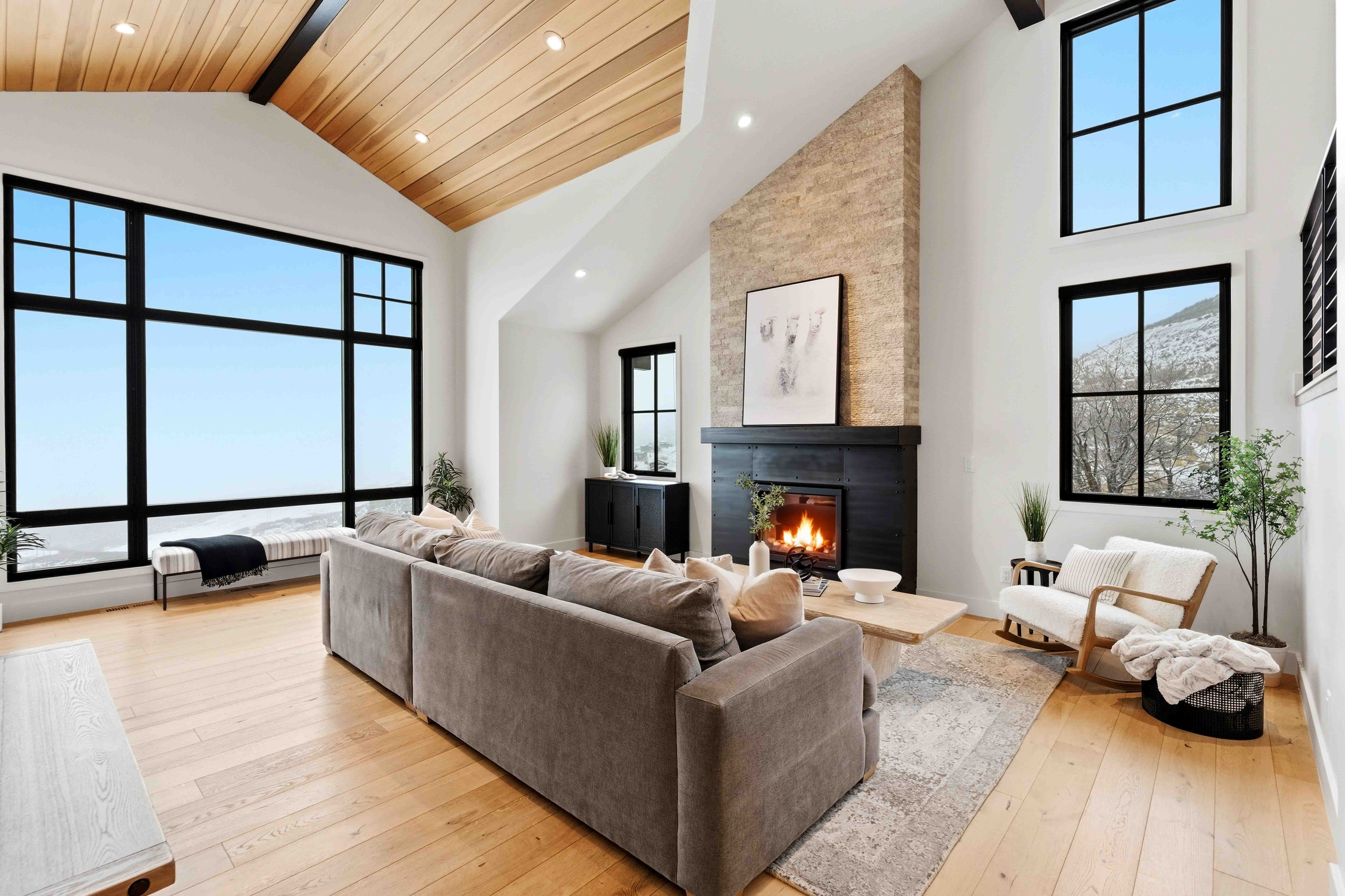 Modern living room with large windows, gray sofa, lit fireplace, wooden ceiling, and light hardwood floor.