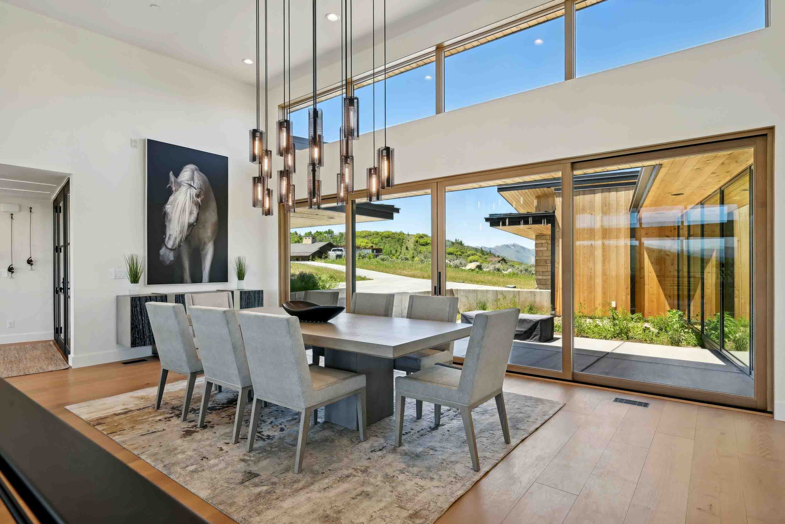 Modern dining room with a large table, eight gray chairs, hanging lights, and large glass sliding doors.