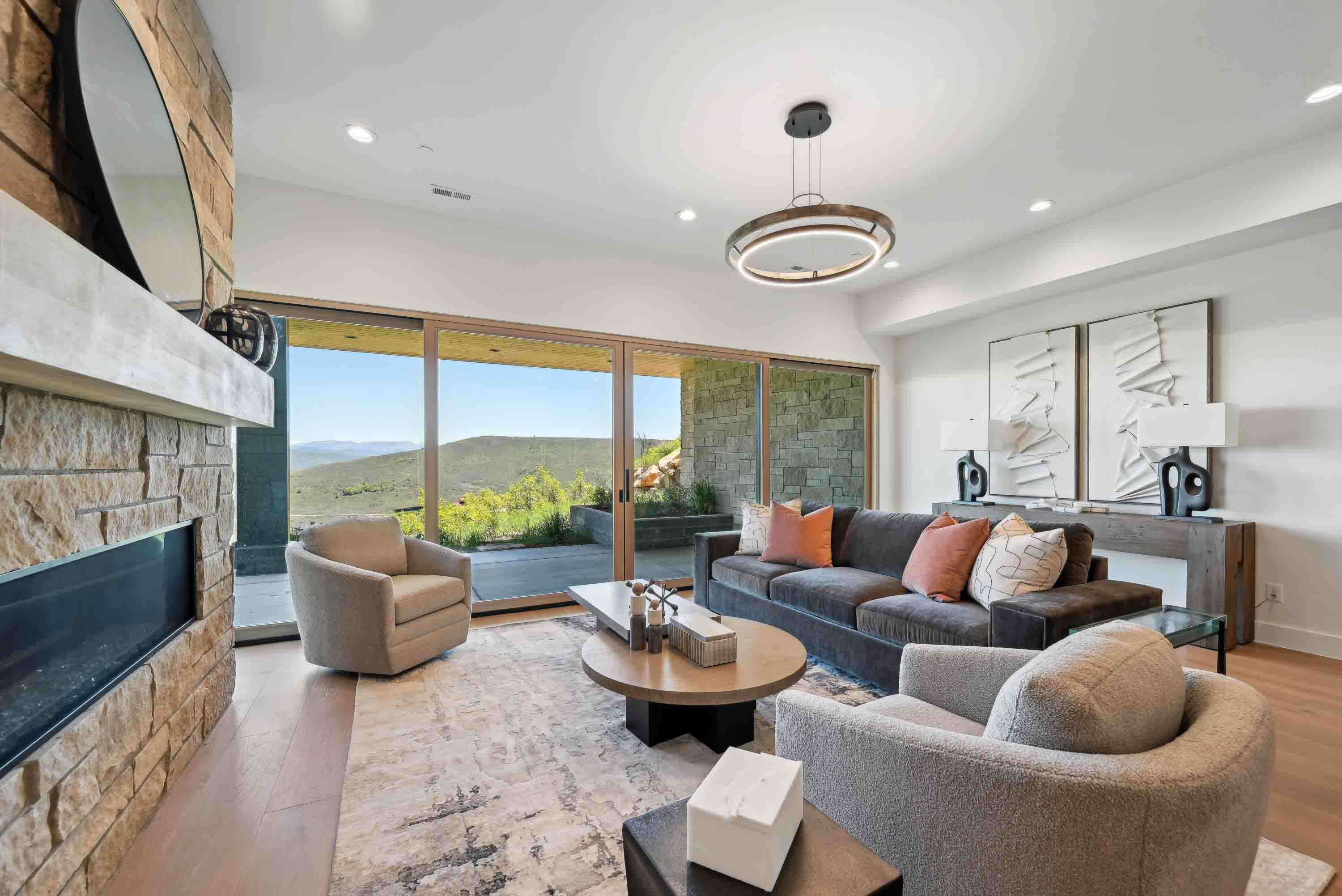 Modern living room with stone fireplace, large windows, gray sofa with pillows, and two armchairs.