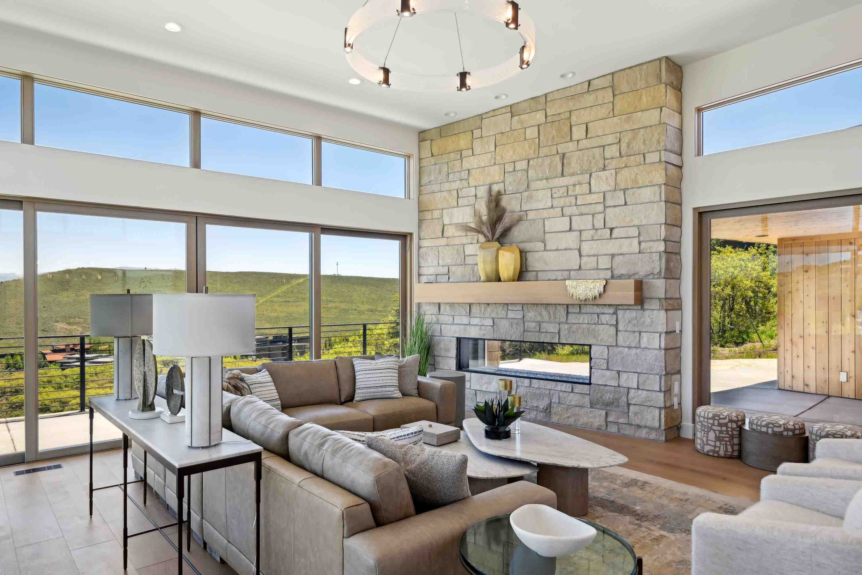 Modern living room with stone fireplace, large windows overlooking green hills, beige sofas, and a coffee table.