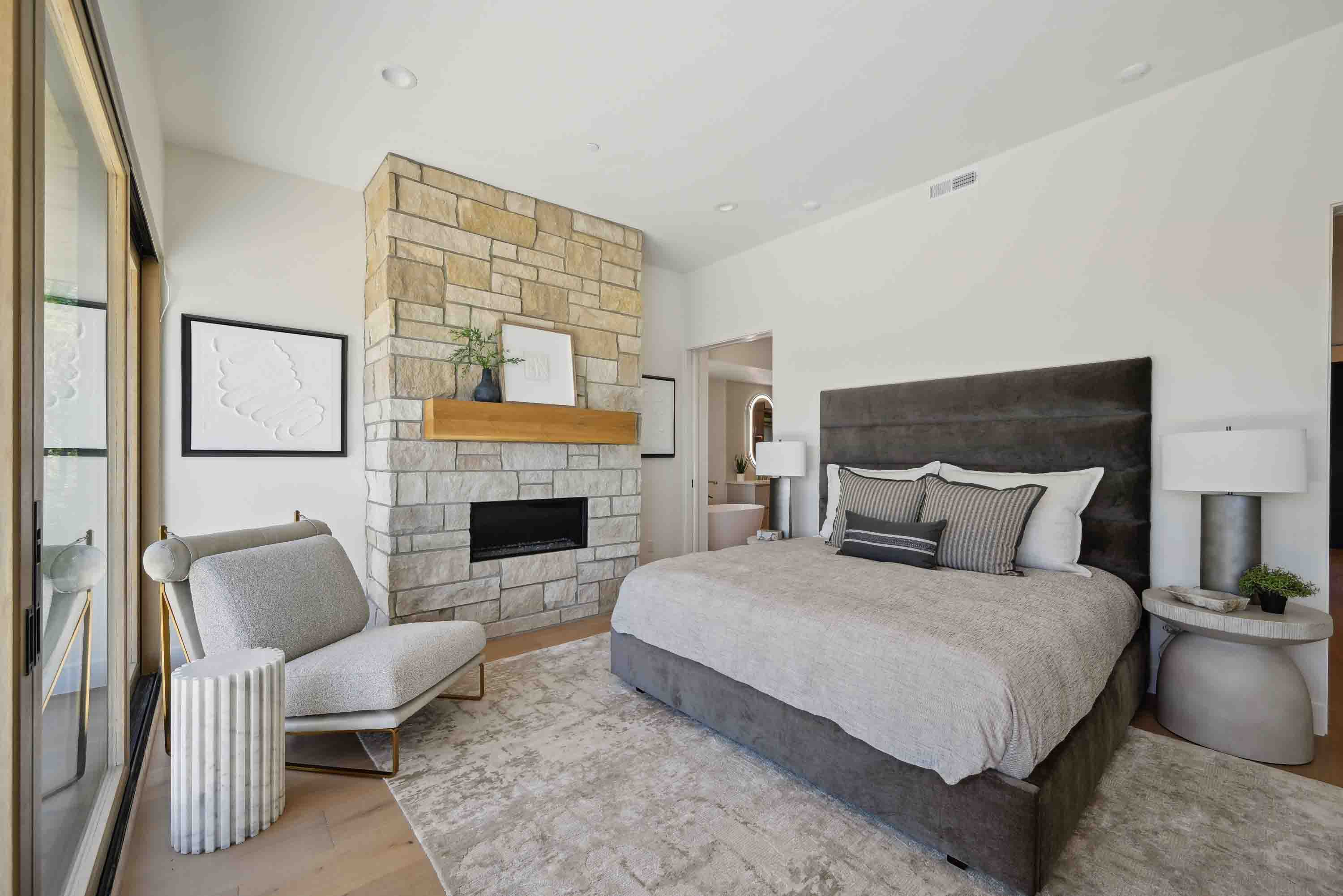 Modern bedroom with stone fireplace, gray bed, armchair, and round side table under white walls.