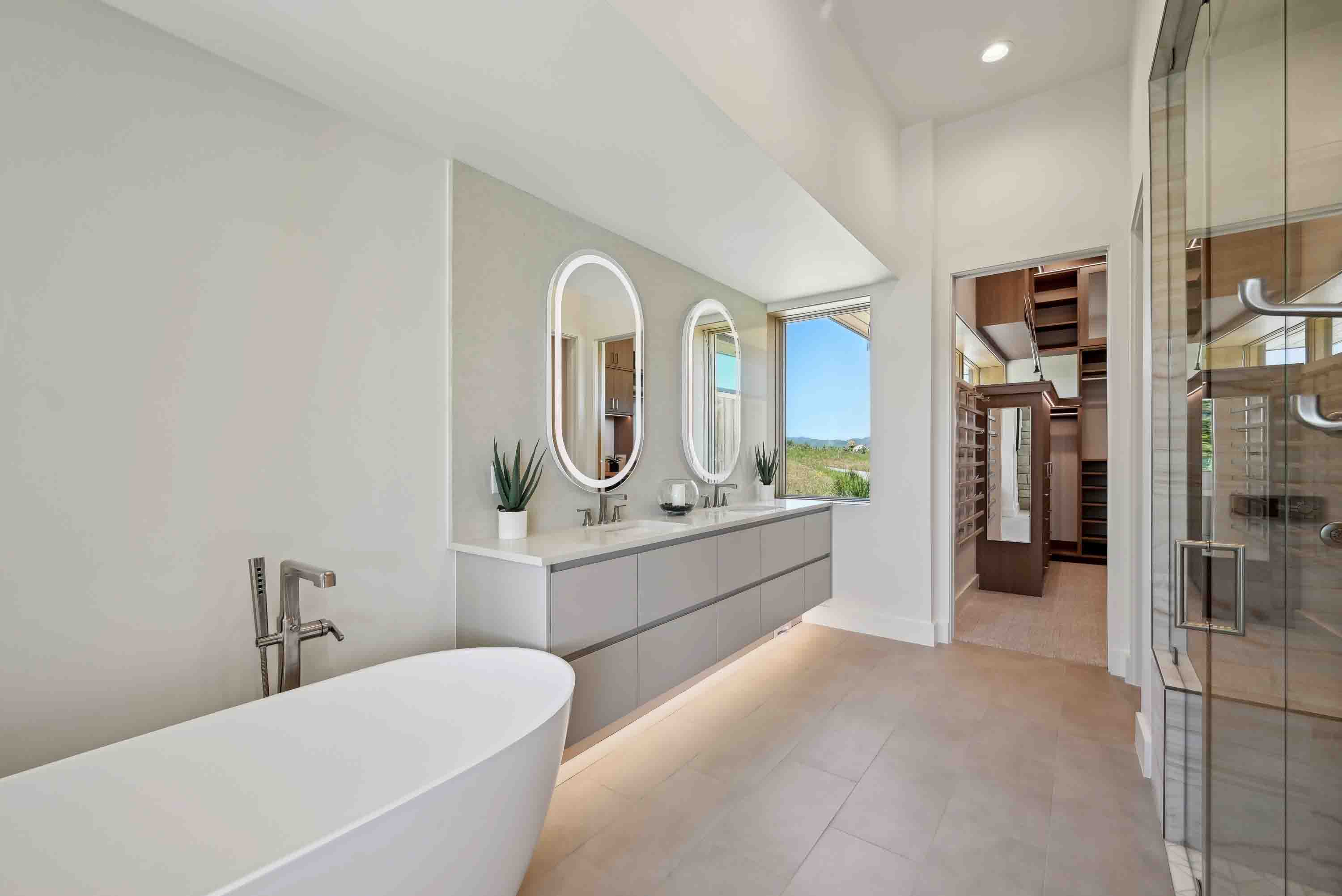 Modern bathroom with freestanding tub, double sinks with backlit mirrors, and walk-in closet visible.