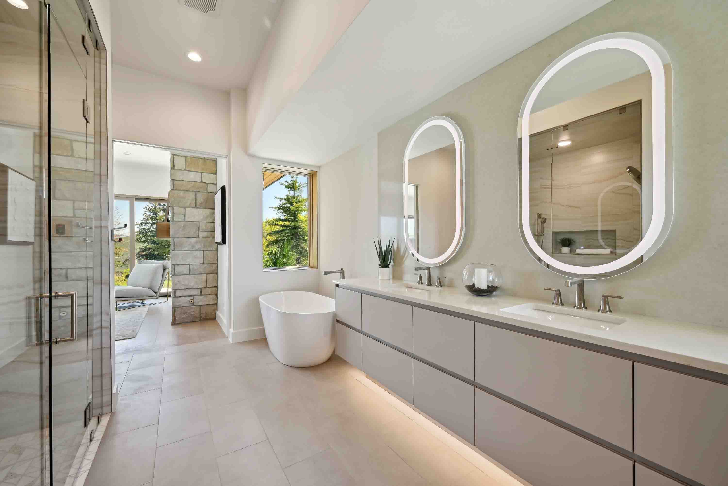 Modern bathroom with freestanding tub, double sinks, lighted mirrors, and glass shower door.