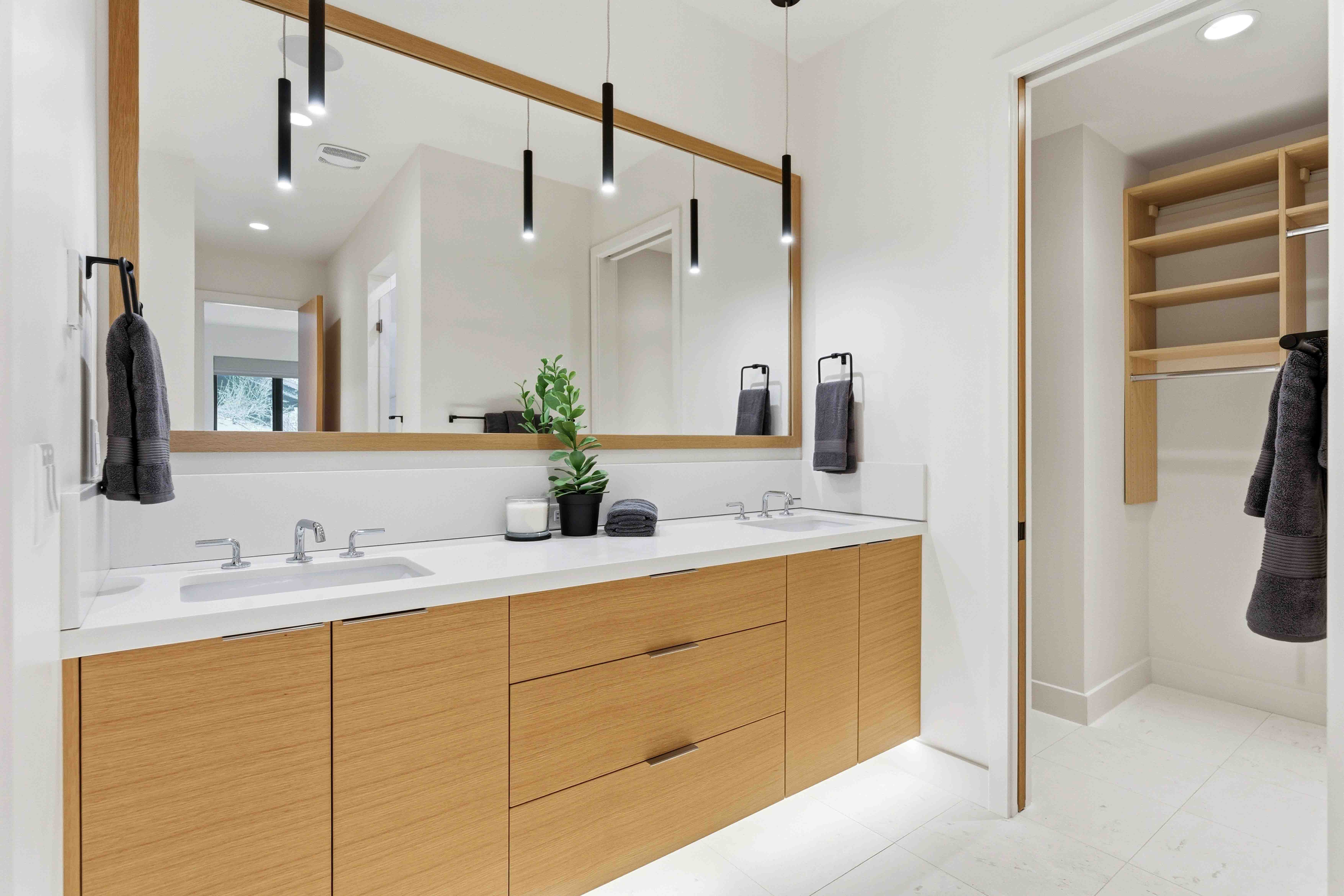 Modern bathroom with double sinks, wooden cabinetry, large mirror, black towels, and a walk-in closet.