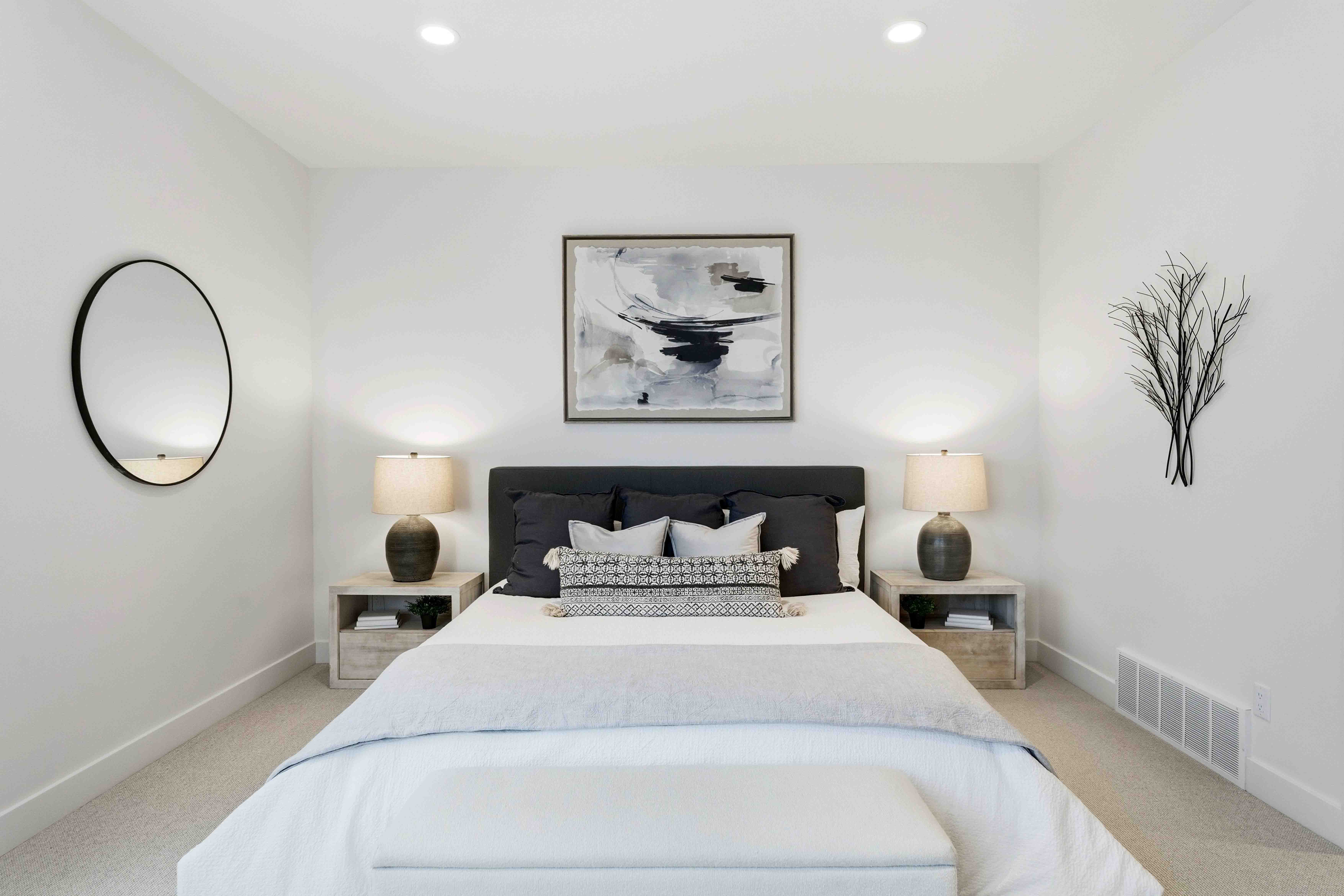 Modern bedroom with bed, black and gray pillows, two lamps on wooden nightstands, round mirror, and wall art.