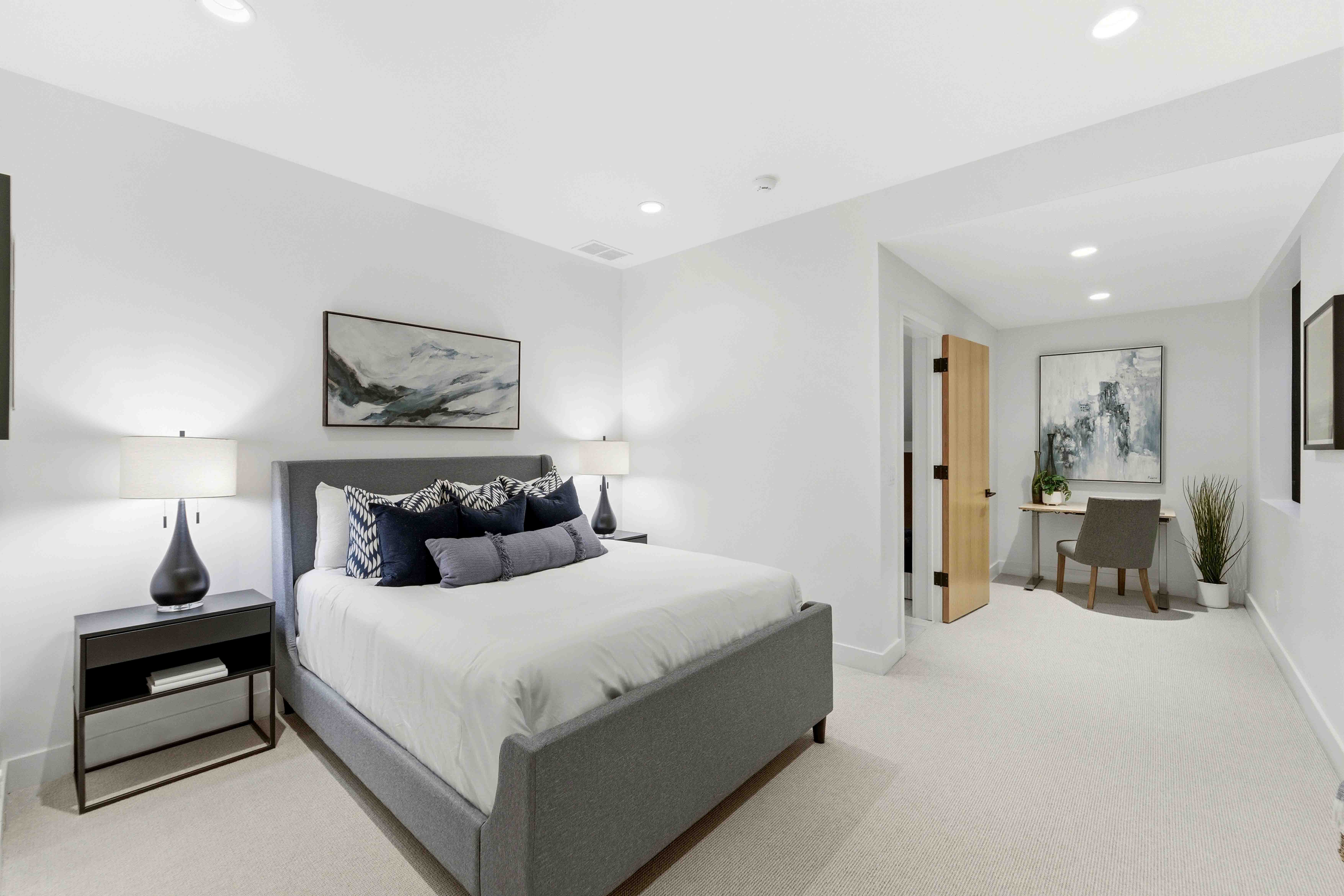 Modern bedroom with gray upholstered bed, two bedside lamps, and a small desk in the corner.