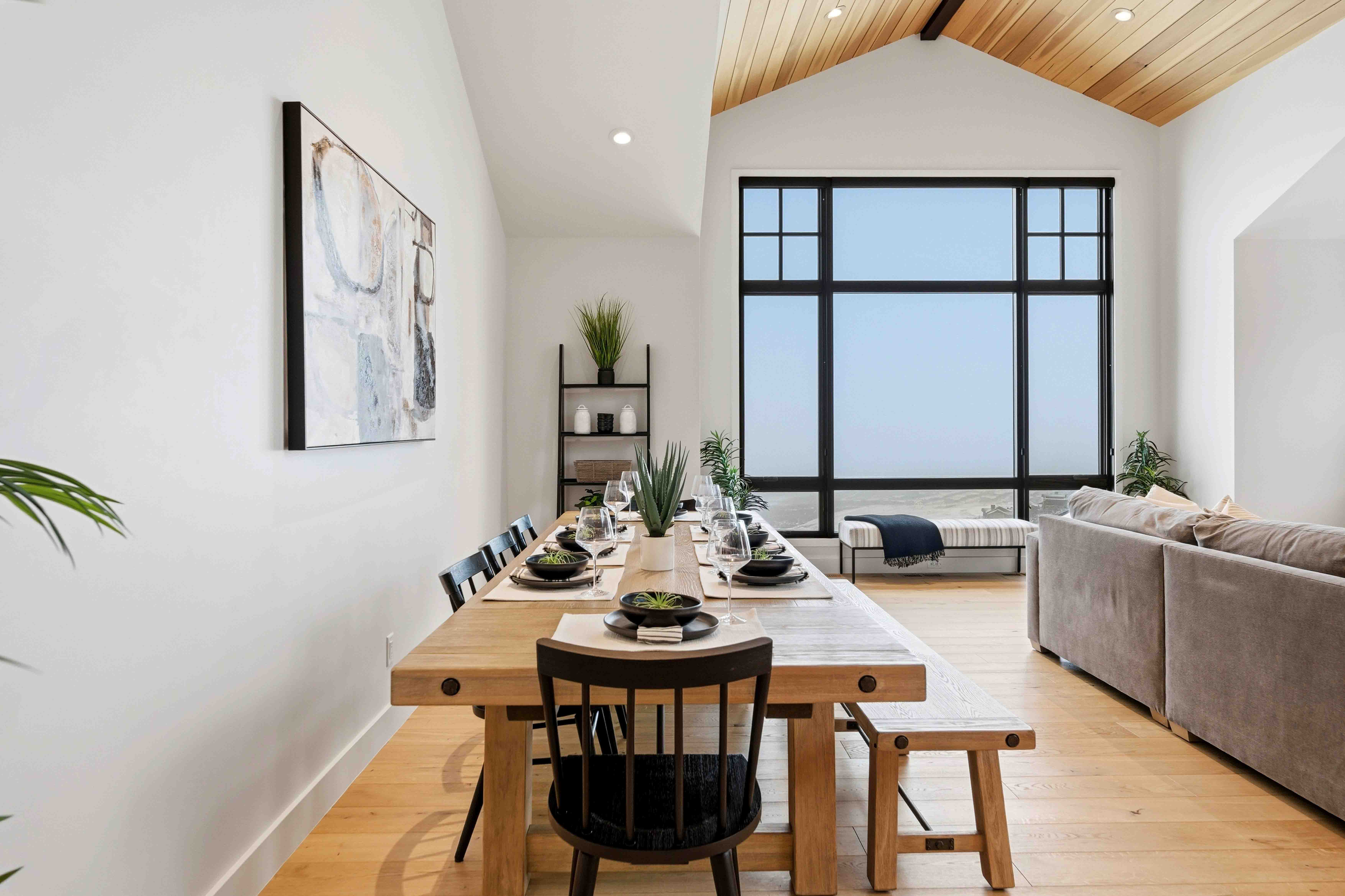 Modern dining room with wooden table, black chairs, large window, gray couch, and wooden ceiling.