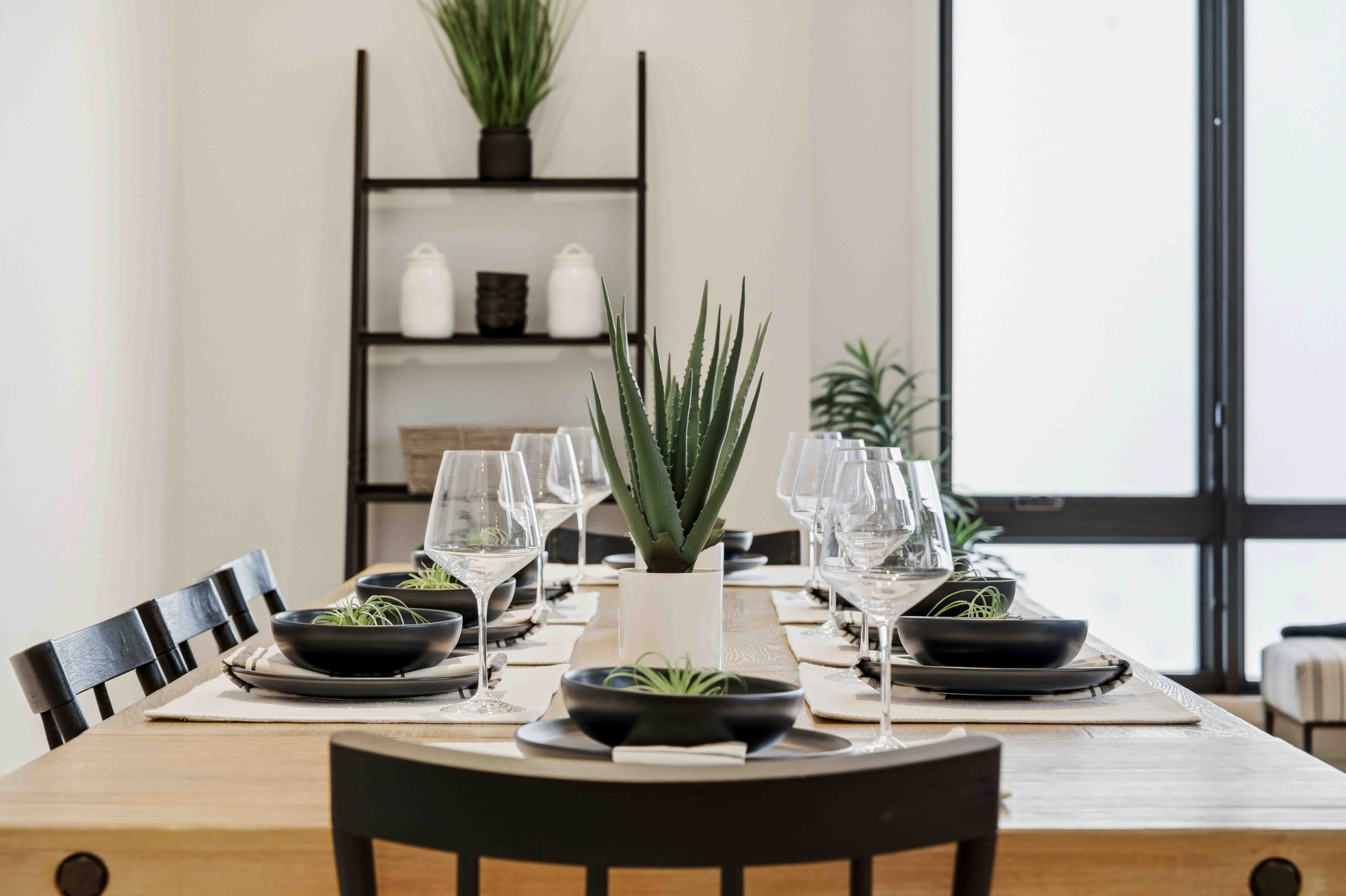 Modern dining table set with black bowls, wine glasses, and green succulent plants in a bright room.
