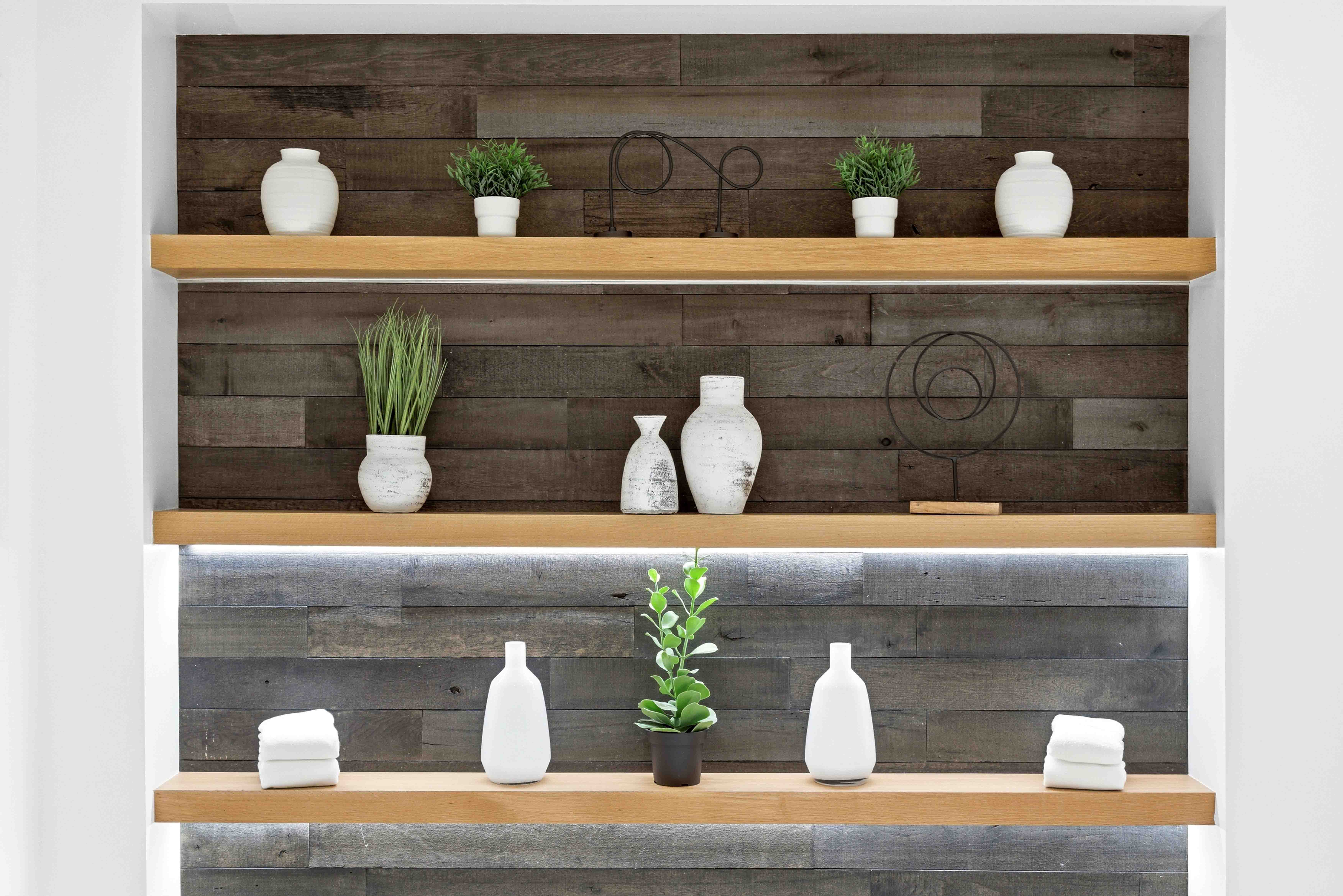 Three wooden shelves on a dark wood wall hold white vases, green plants, sculptures, and folded white towels.