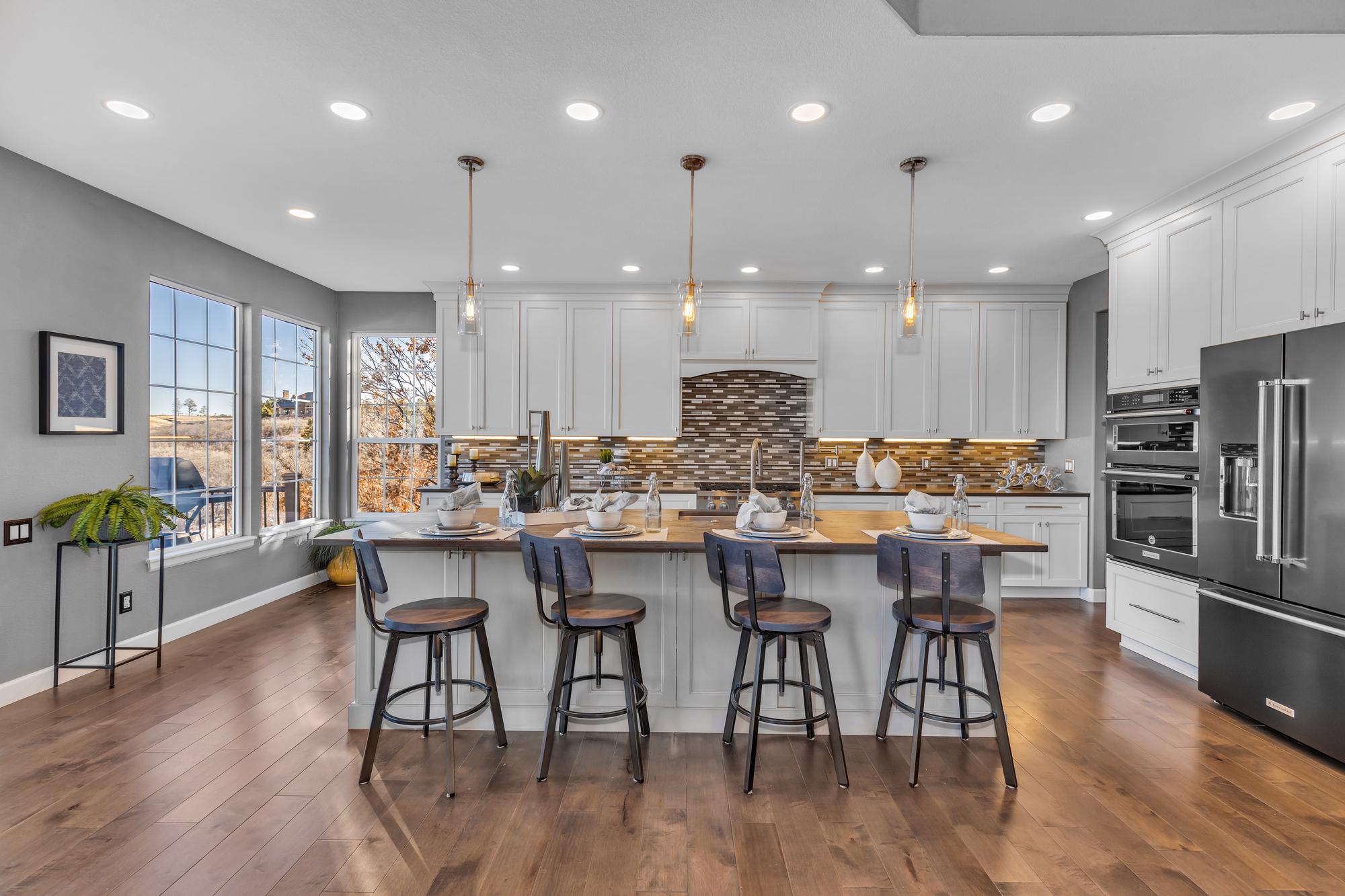 Modern kitchen with white cabinets, wooden island with four stools, and large windows showing outdoor view.