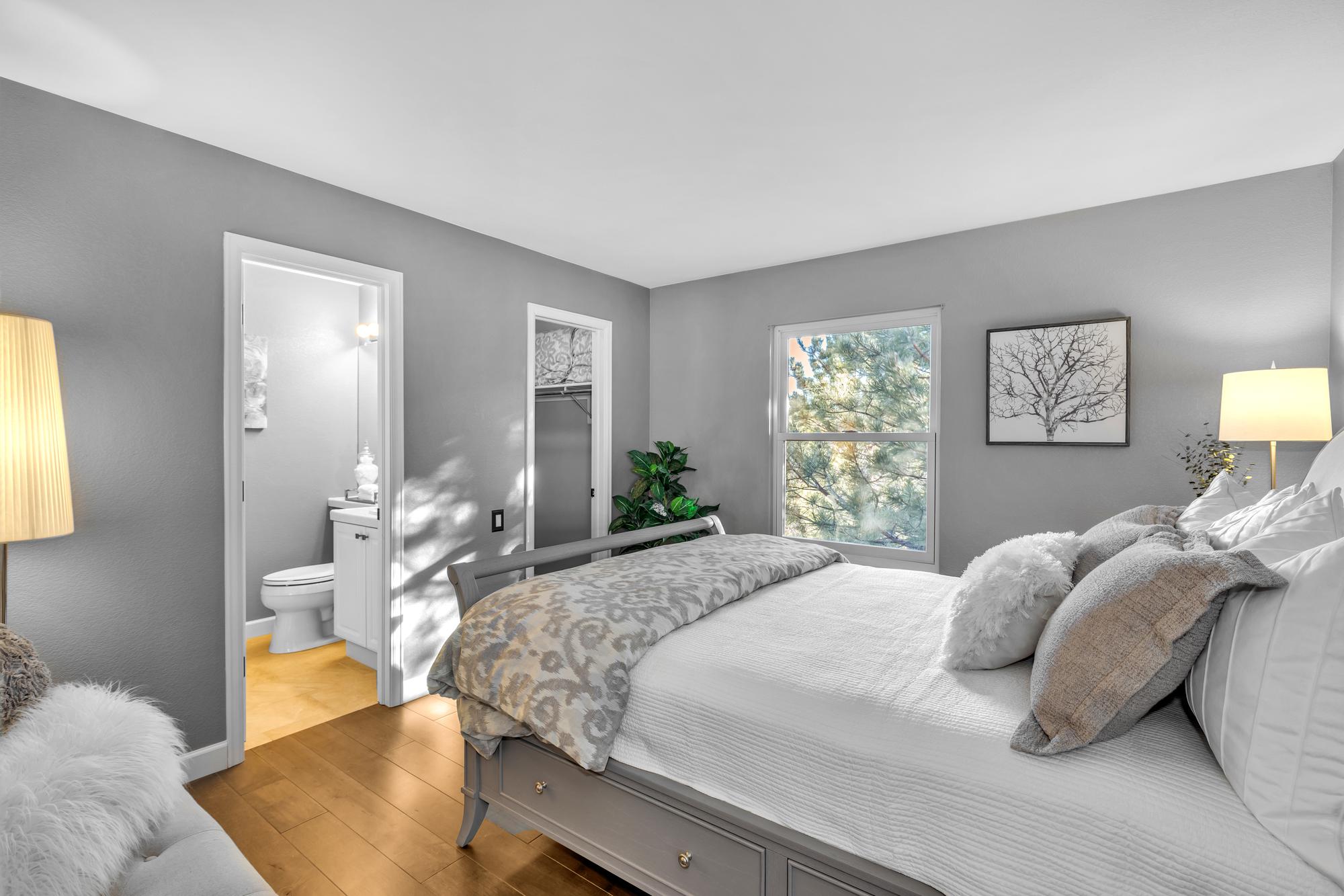 Cozy bedroom with gray walls, a bed with white and beige bedding, lamps, and a window showing green trees.
