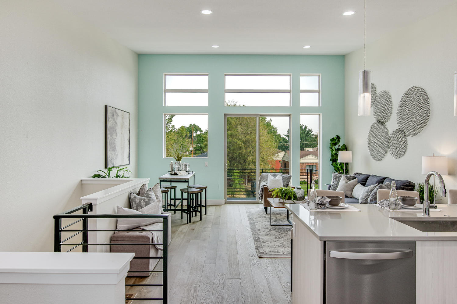Bright living room with teal accent wall, large windows, dining table, sofas, and kitchen island with sink.