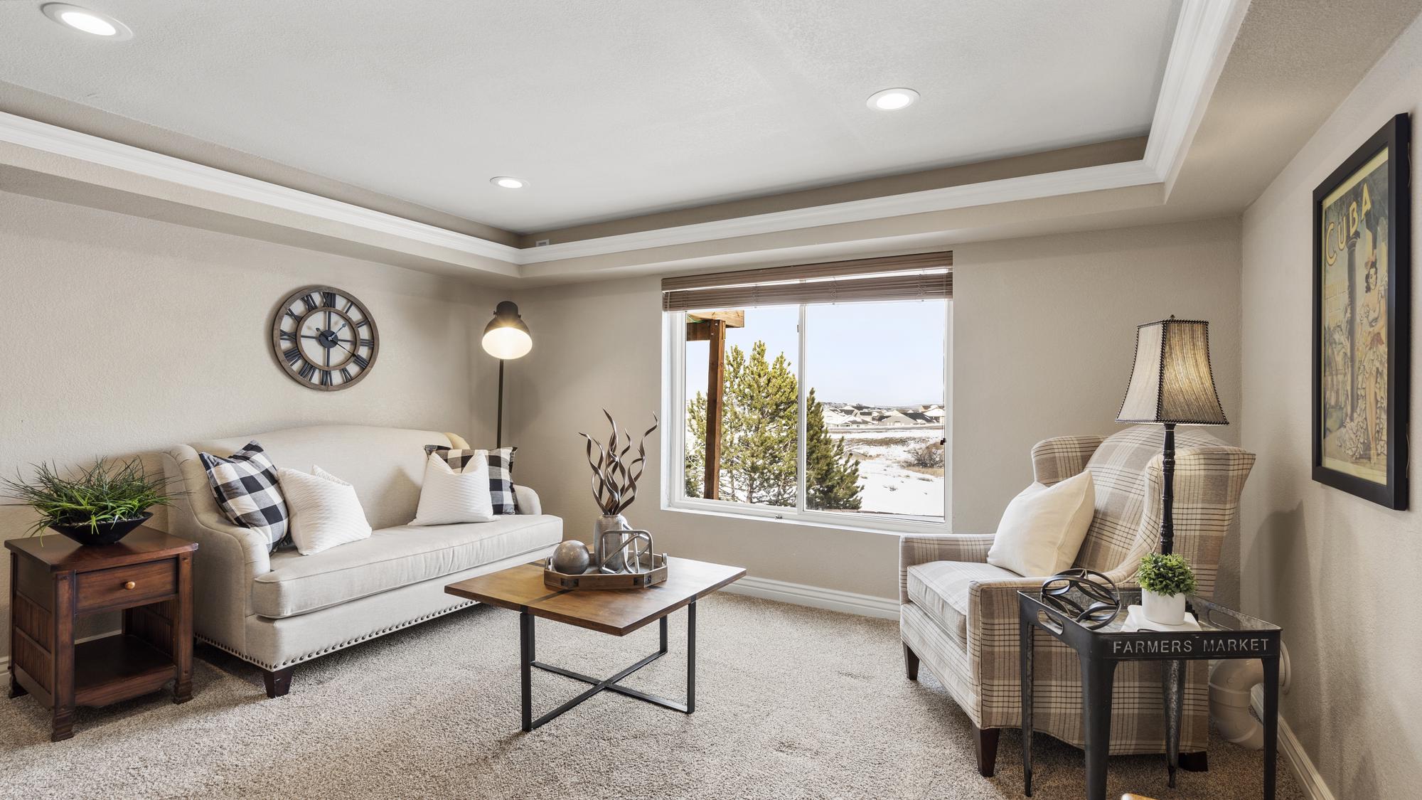 Cozy living room with beige sofa, plaid armchair, wooden coffee table, and a window showing trees outside.