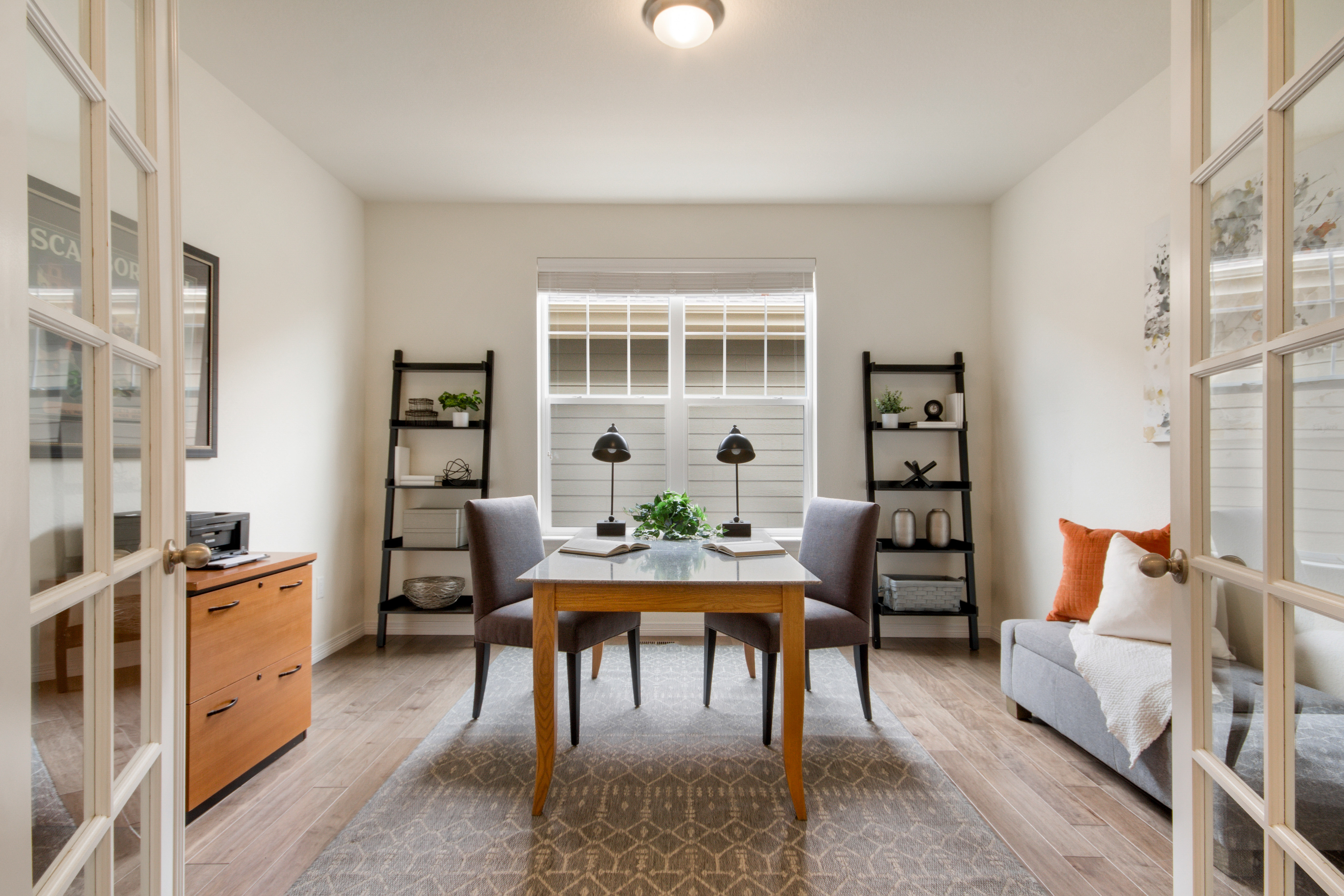 Modern home office with wooden table, two chairs, decorative shelves, and sofa with pillows.