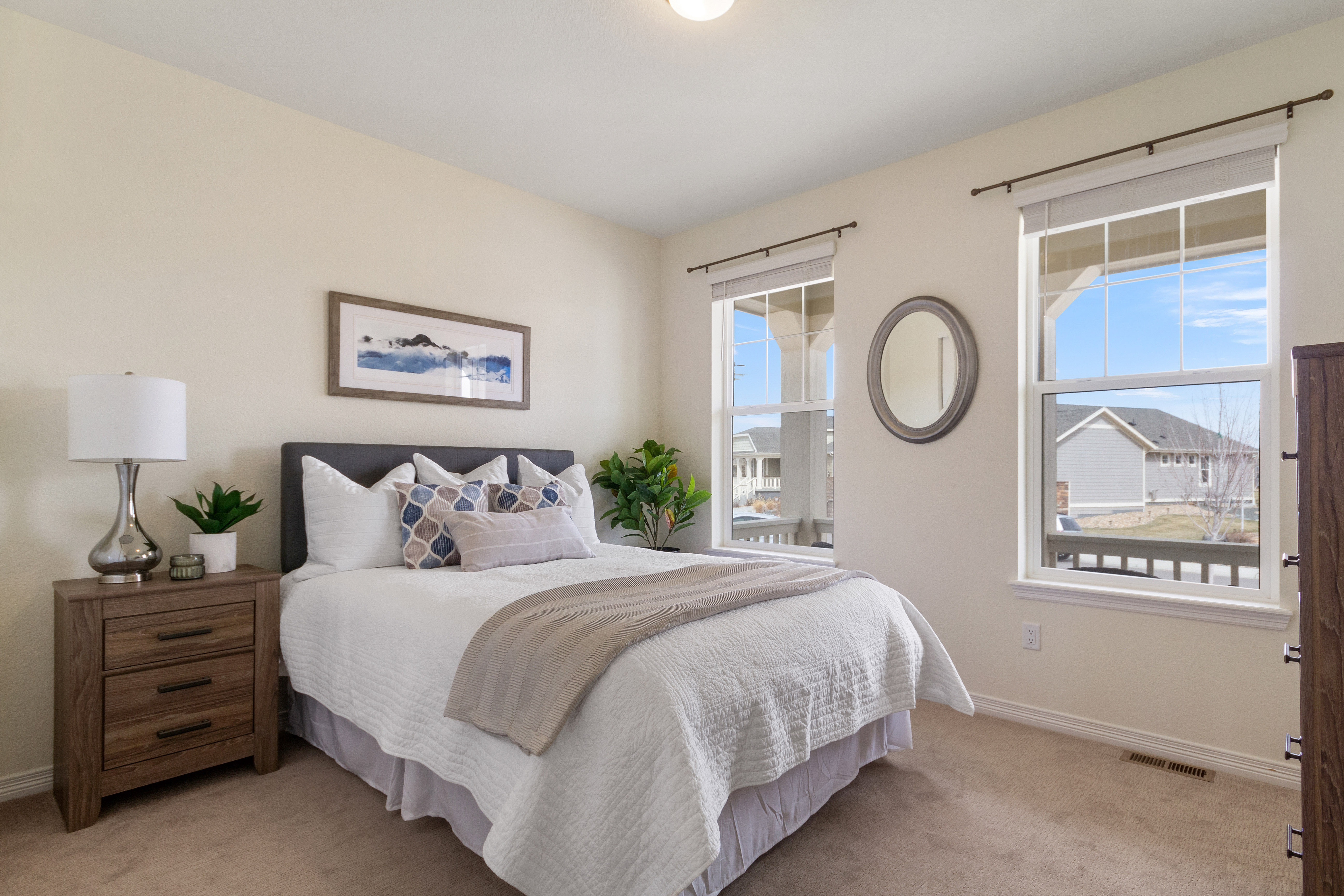 Bright bedroom with a neatly made bed, wooden nightstand, two large windows, and a round wall mirror.