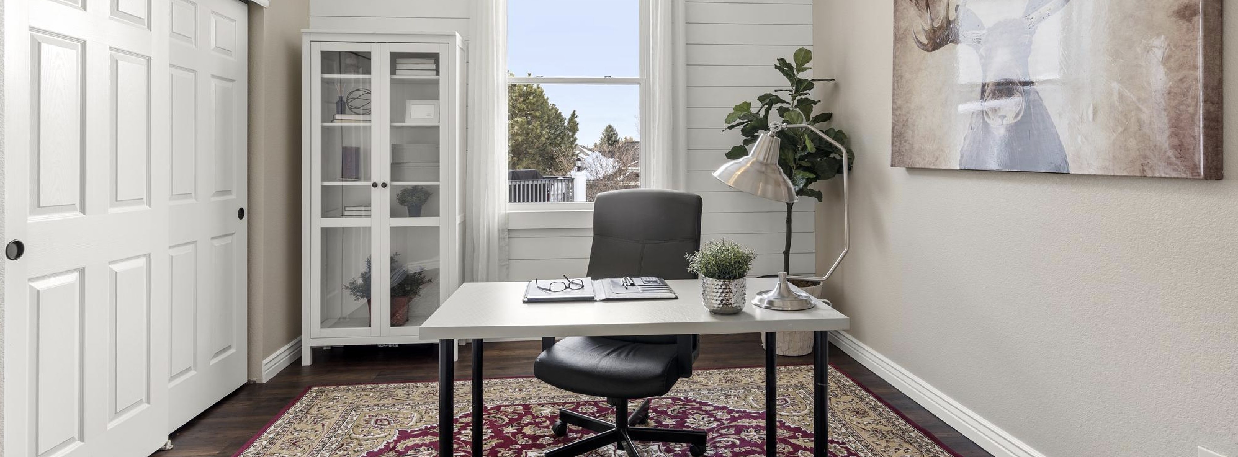 Minimalist home office with desk, black chair, lamp, plant, and glass cabinet near a window.