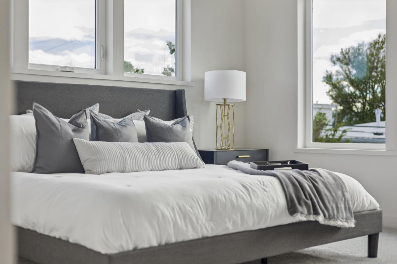 Modern bedroom with a grey upholstered bed, white bedding, grey pillows, and a black nightstand with a lamp.
