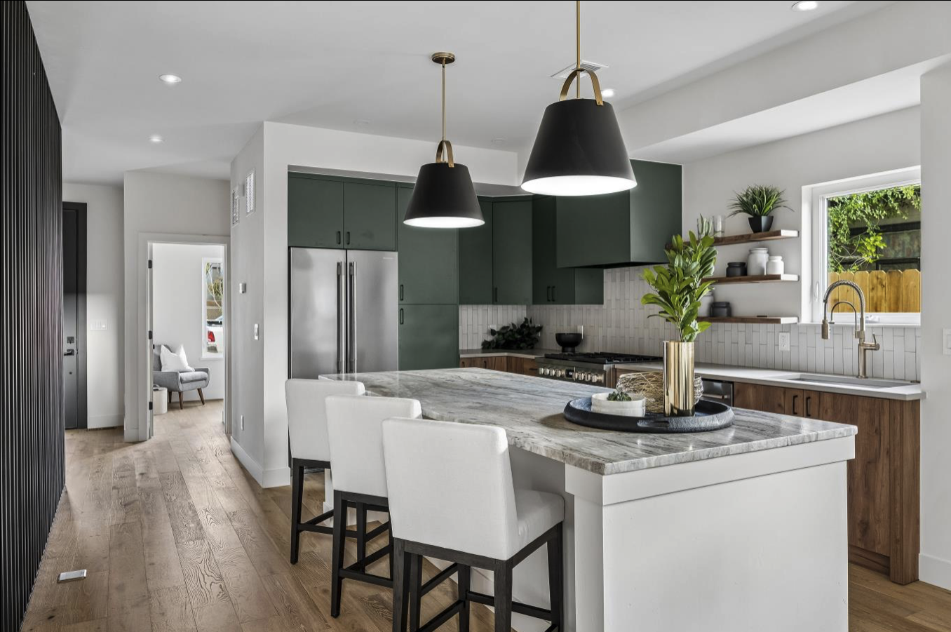 Modern kitchen with marble island, white chairs, green cabinets, wooden shelves, and black pendant lights.