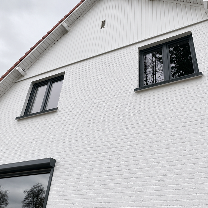 Façade de maison en briques blanches avec fenêtres à encadrement noir et toit de tuiles sous un ciel nuageux.