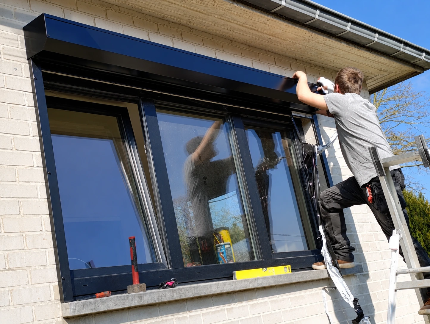 Un ouvrier sur une échelle installe un auvent métallique noir au-dessus d'une fenêtre à cadre noir sur un bâtiment en briques blanches.