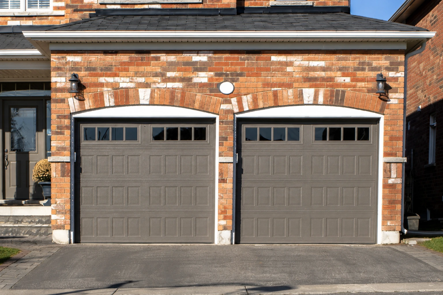 Deux portes de garage marron fermées, encastrées dans une maison en briques rouges, avec des éclairages extérieurs au-dessus de chaque porte.