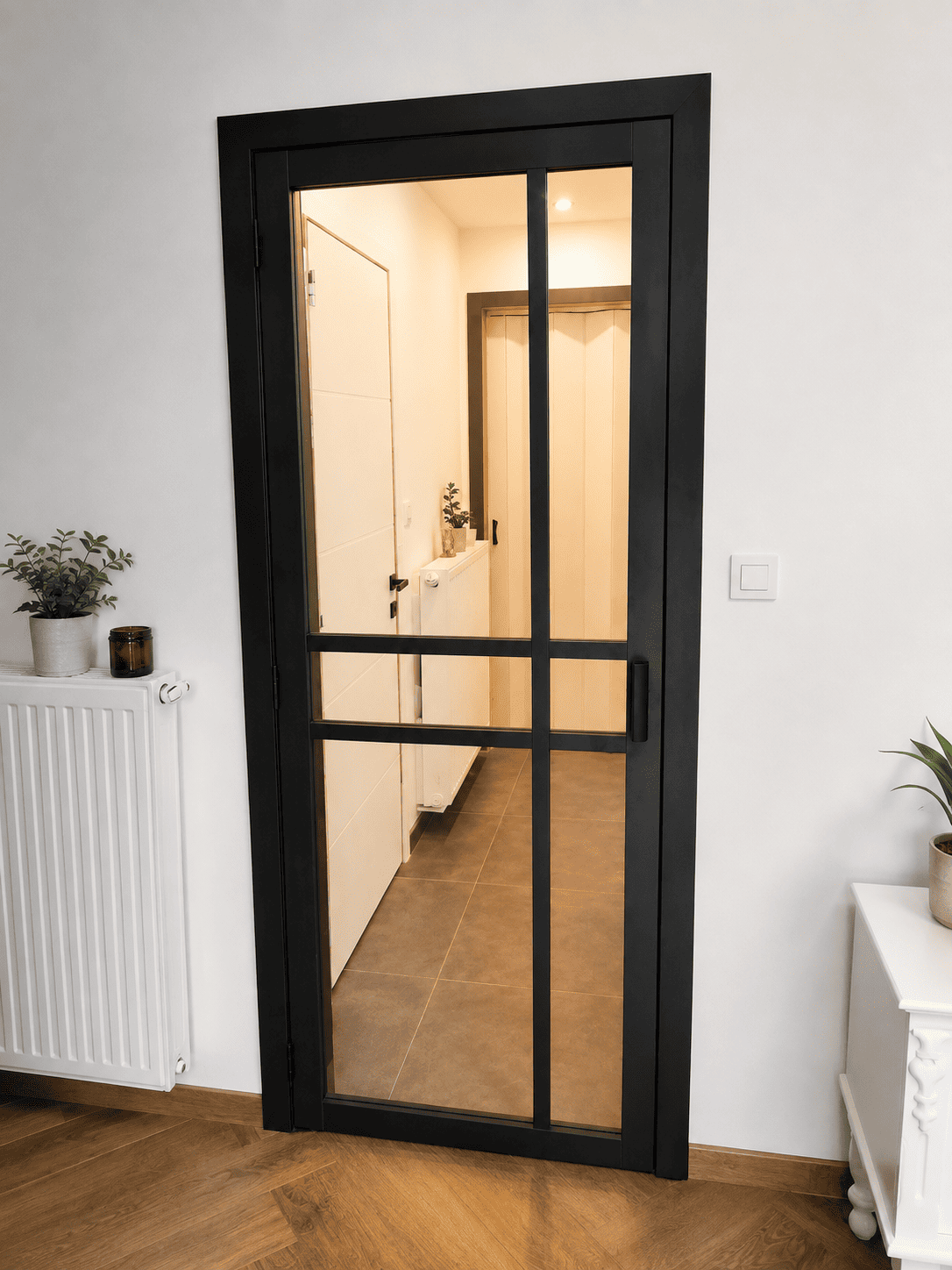 Porte intérieure vitrée noire avec cadre métallique et poignée, sol parquet et décor minimaliste.
