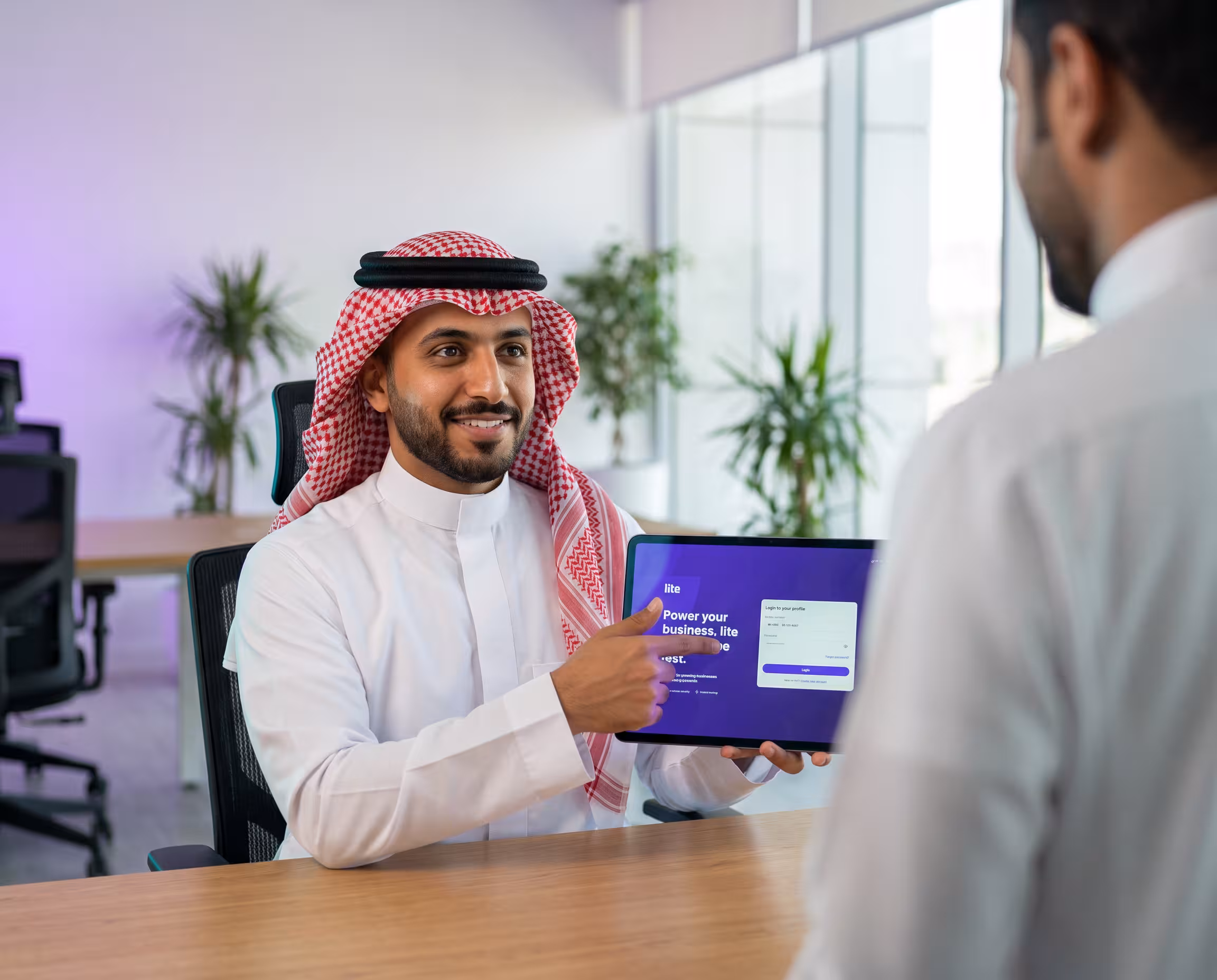 Smiling man in checkered keffiyeh and white thobe holding card, paying at a terminal.