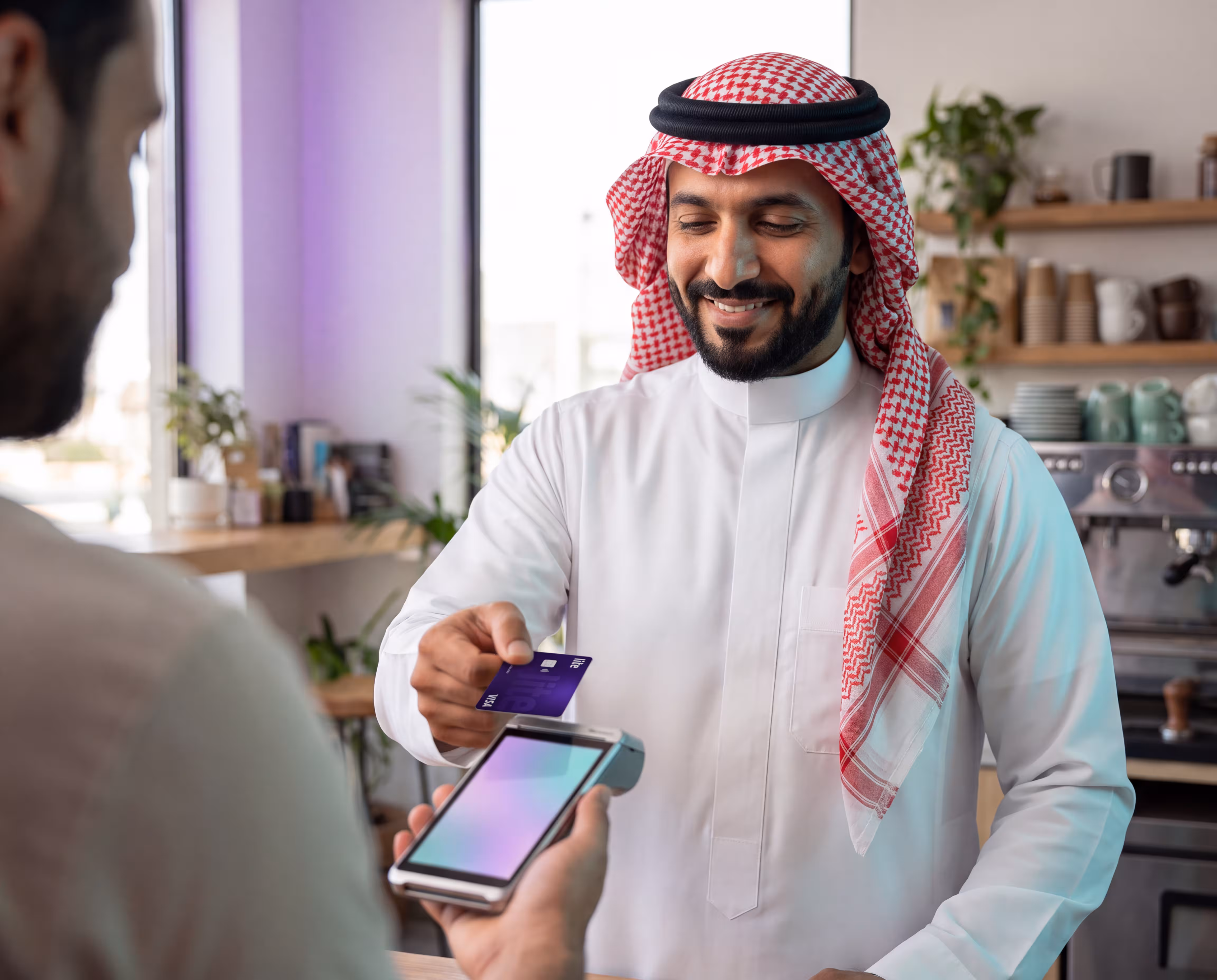 Smiling man in checkered keffiyeh and white thobe holding card, paying at a terminal.