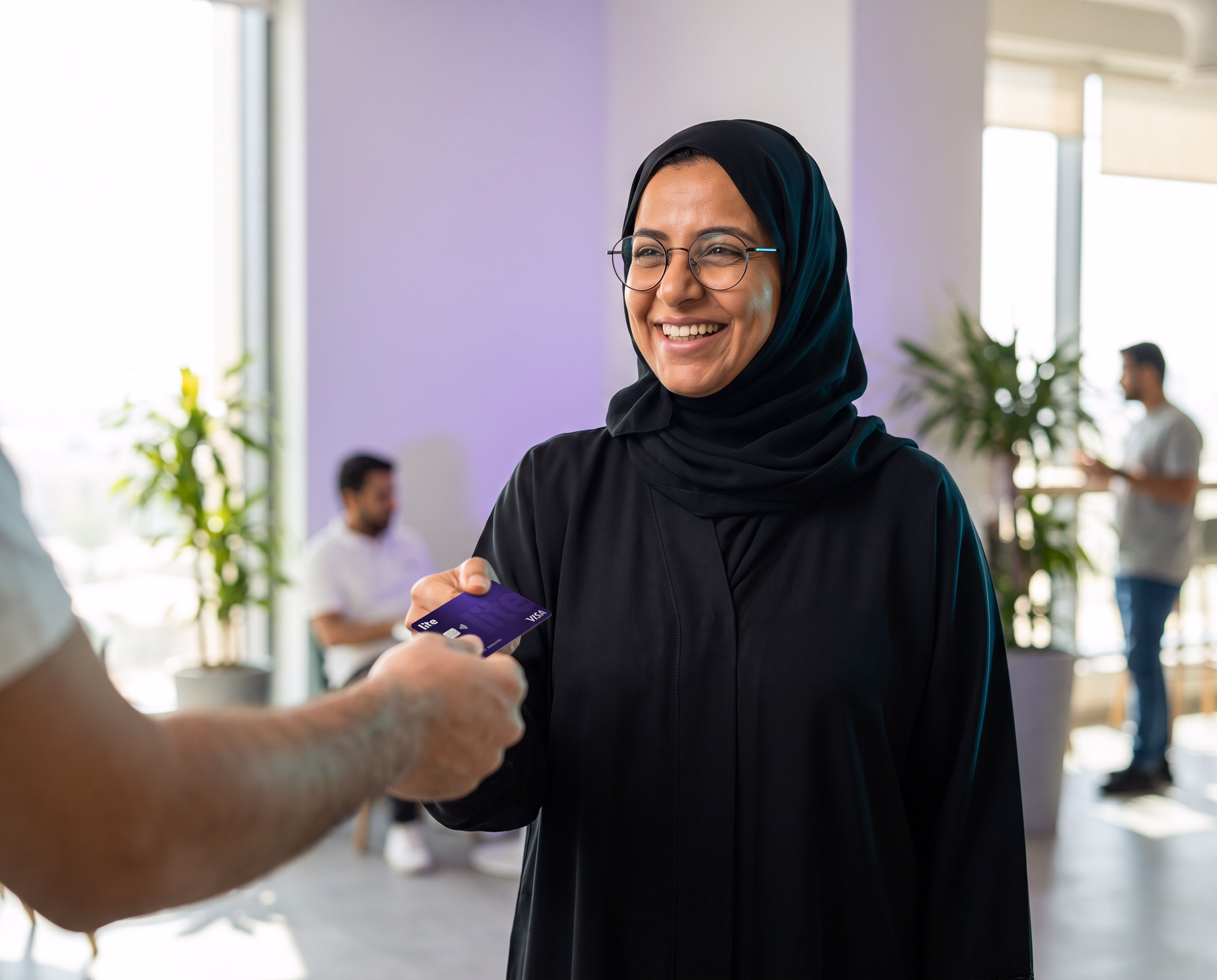 Smiling man in checkered keffiyeh and white thobe holding card, paying at a terminal.