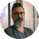 Man with gray beard and glasses wearing a dark gray V-neck shirt standing indoors.