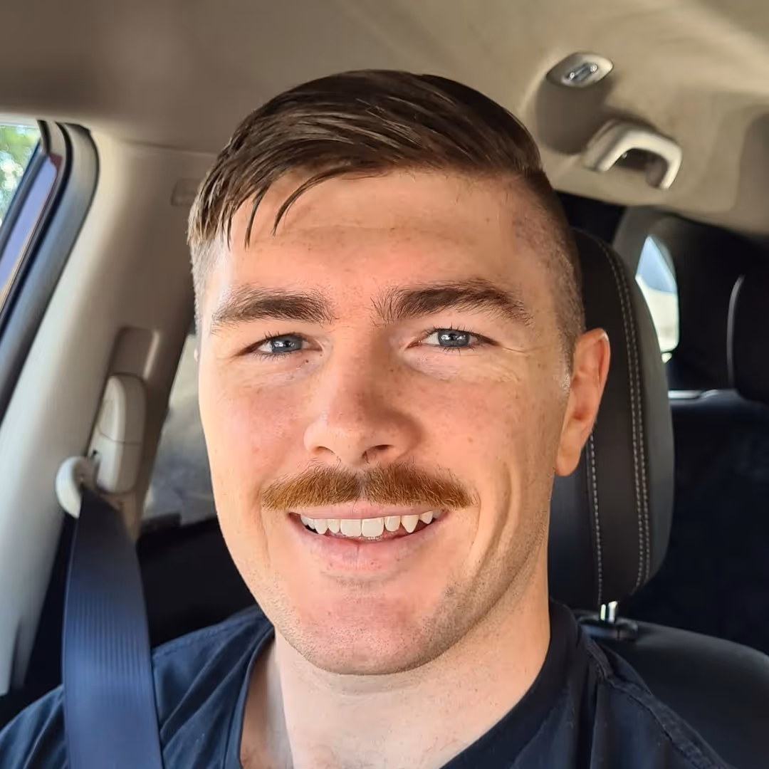 Smiling man with short brown hair and a mustache wearing a black shirt and seatbelt in a car.