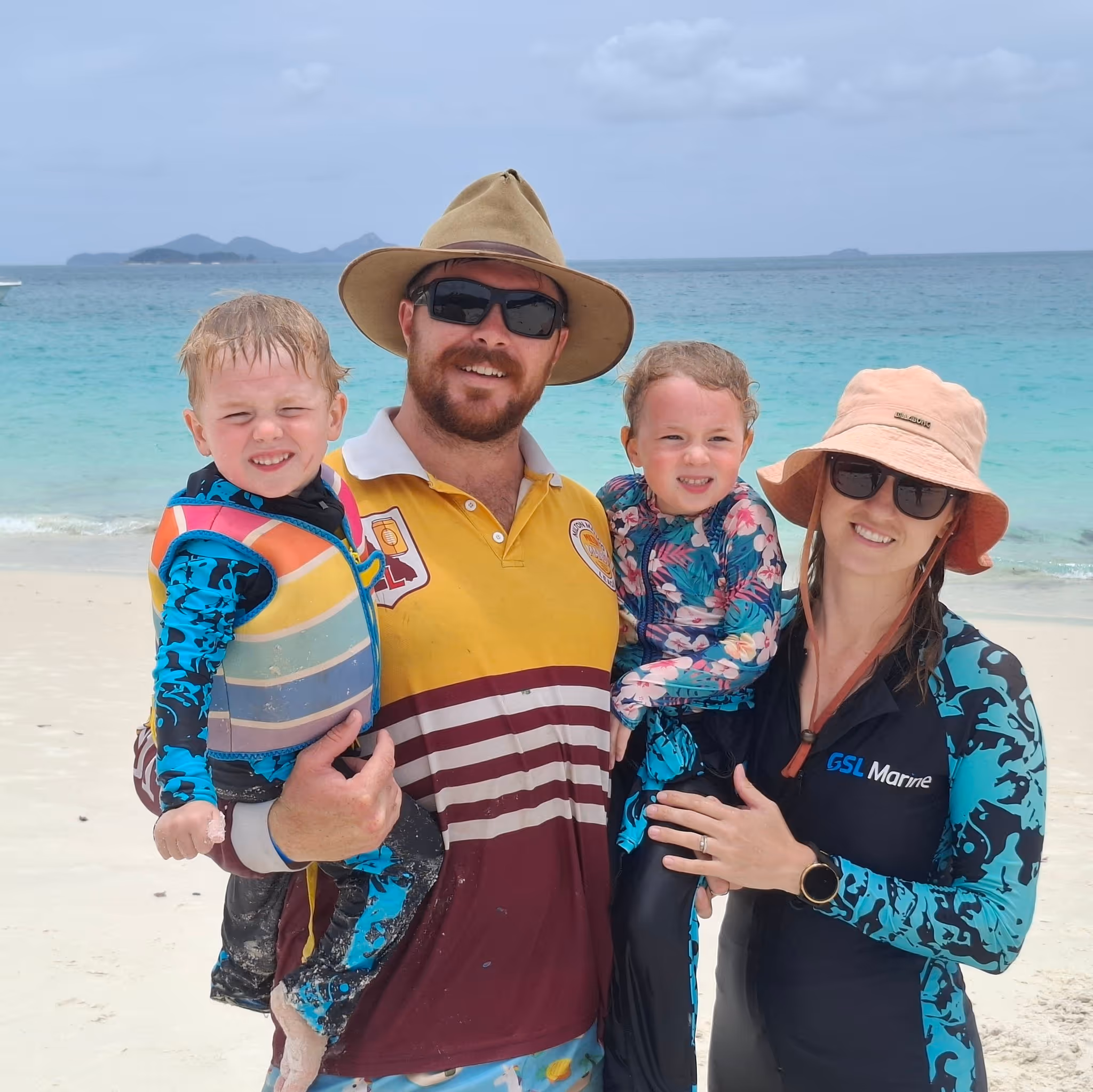 Family of four smiling on a sandy beach with turquoise ocean and distant islands in the background.