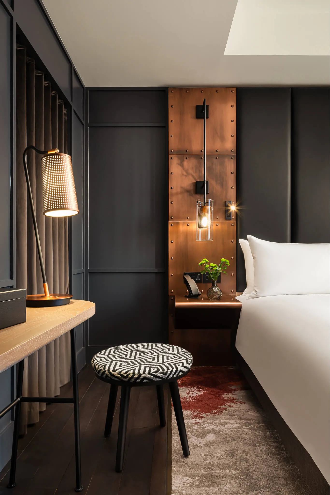 Modern bedroom corner with a wooden desk, patterned round stool, copper wall panel with a hanging light, white bed, and a small vase with green flowers.