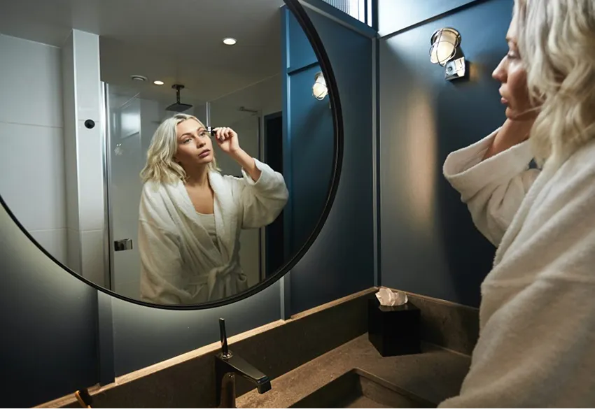 Woman in a white bathrobe applying makeup while looking into a round bathroom mirror.