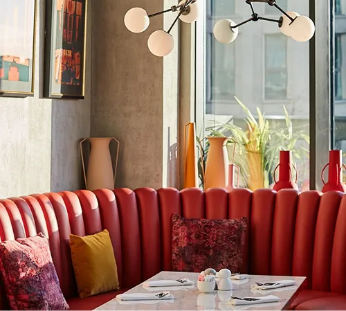 Modern dining booth with red cushioned seating, decorative pillows, marble table set with napkins and cutlery, and large window with tall vases.
