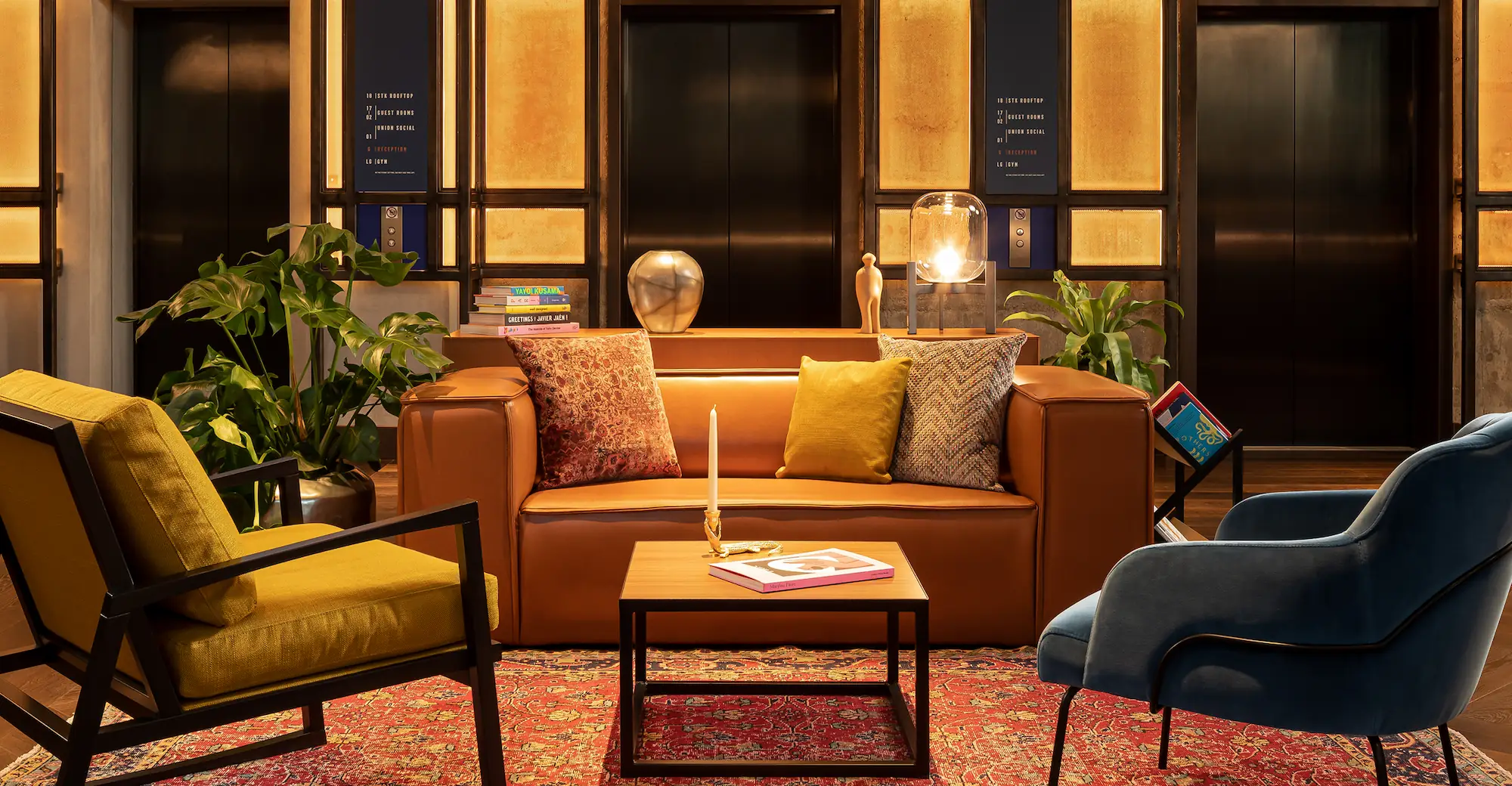 Warmly lit modern living room with a tan leather sofa adorned with three decorative pillows, a yellow armchair, green plants, and a wooden coffee table with a candle and book.