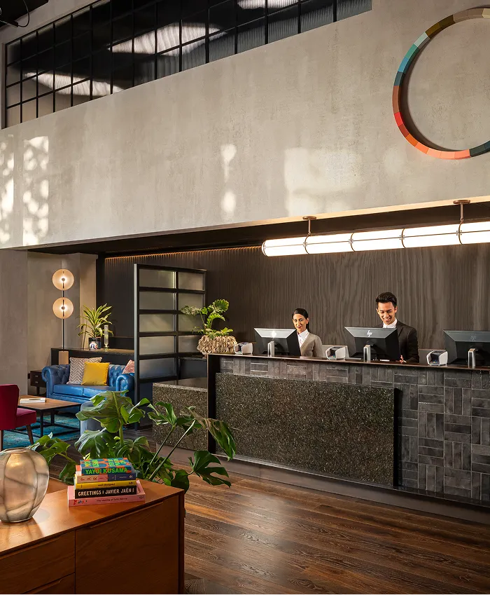 Modern hotel reception area with two receptionists standing behind a dark stone counter, seating area with blue couch, plants, and books on a wooden table.