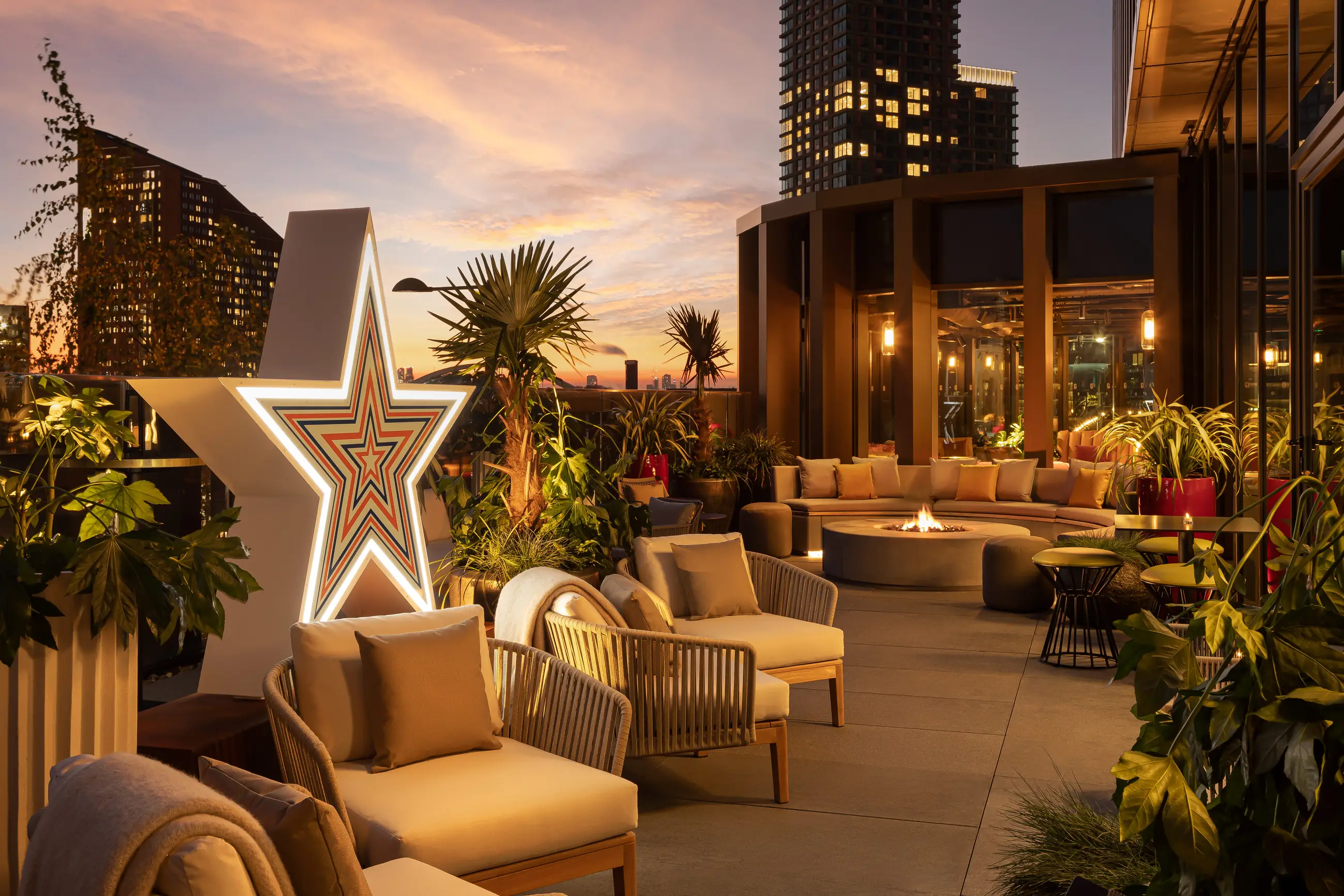 Cozy outdoor terrace at sunset with cushioned chairs, a star-shaped light decoration, plants, and a round fire pit surrounded by a circular sofa.