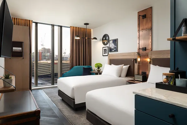 Modern hotel room with two neatly made beds, teal armchair, floor-to-ceiling windows, and city view.