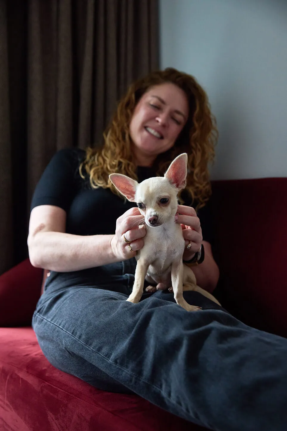 Curly-haired woman sitting on a red sofa holding a small light tan Chihuahua dog on her lap.