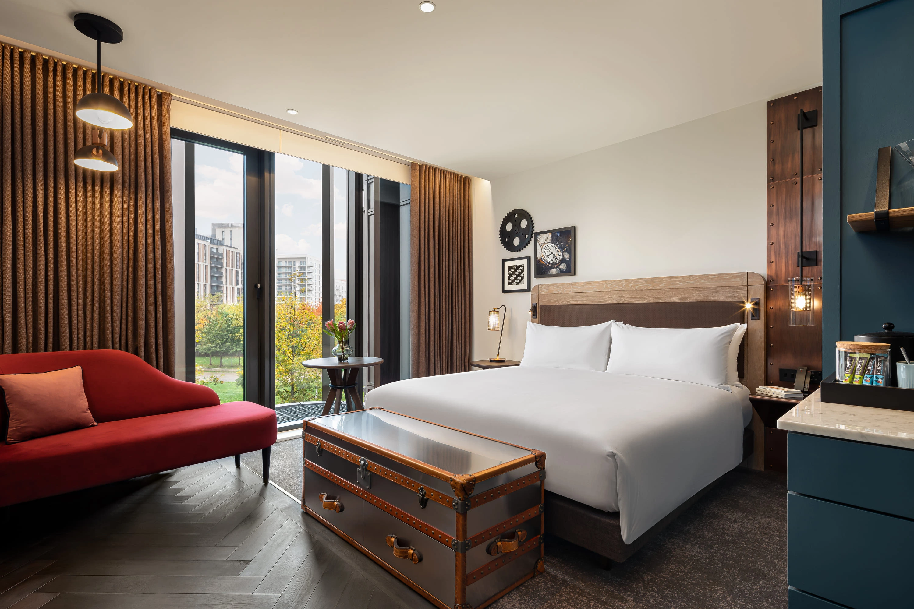 Modern hotel room with large bed, red couch, vintage trunk, and tall windows with city view.
