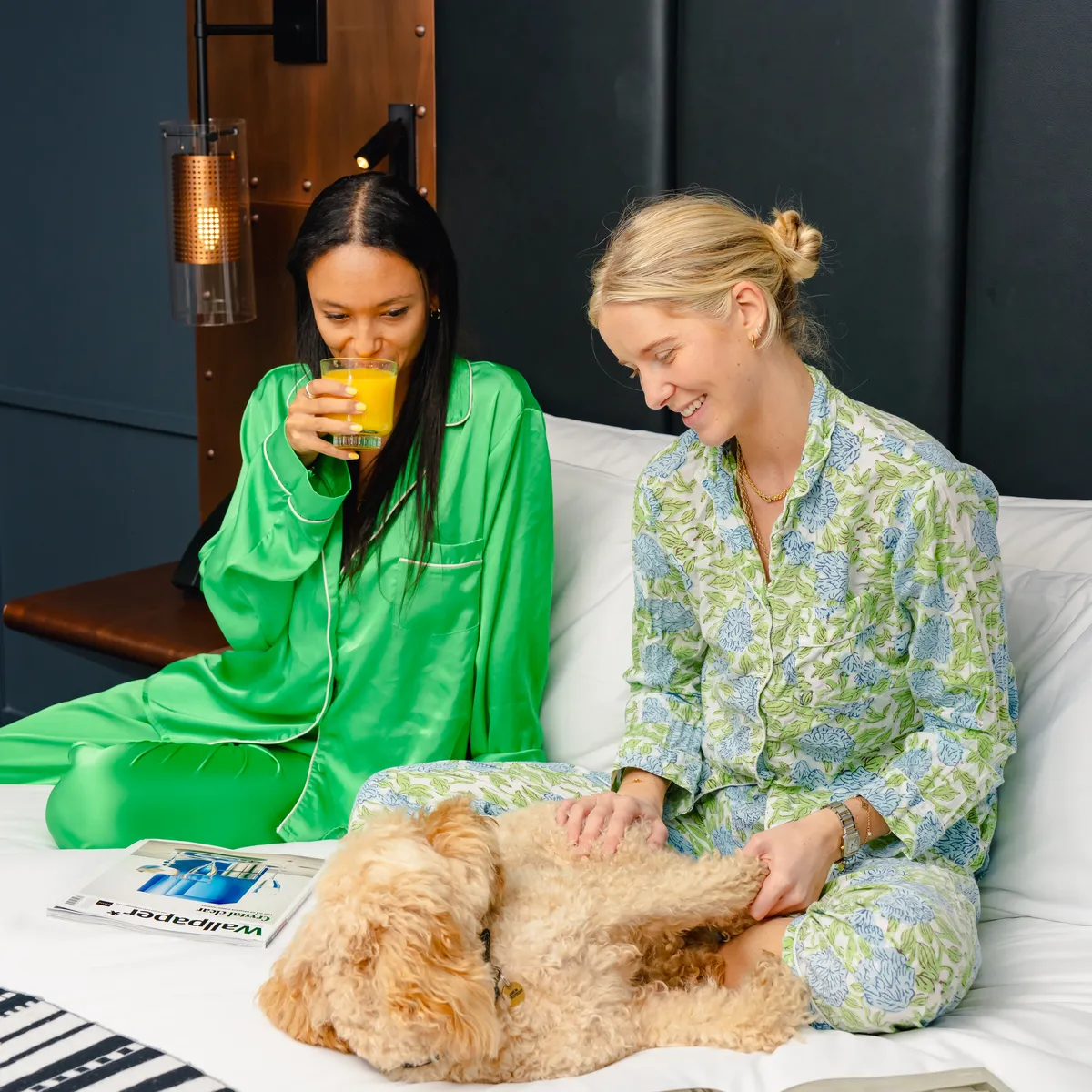 Two women in pajamas relaxing on a bed with a fluffy dog, one drinking orange juice.