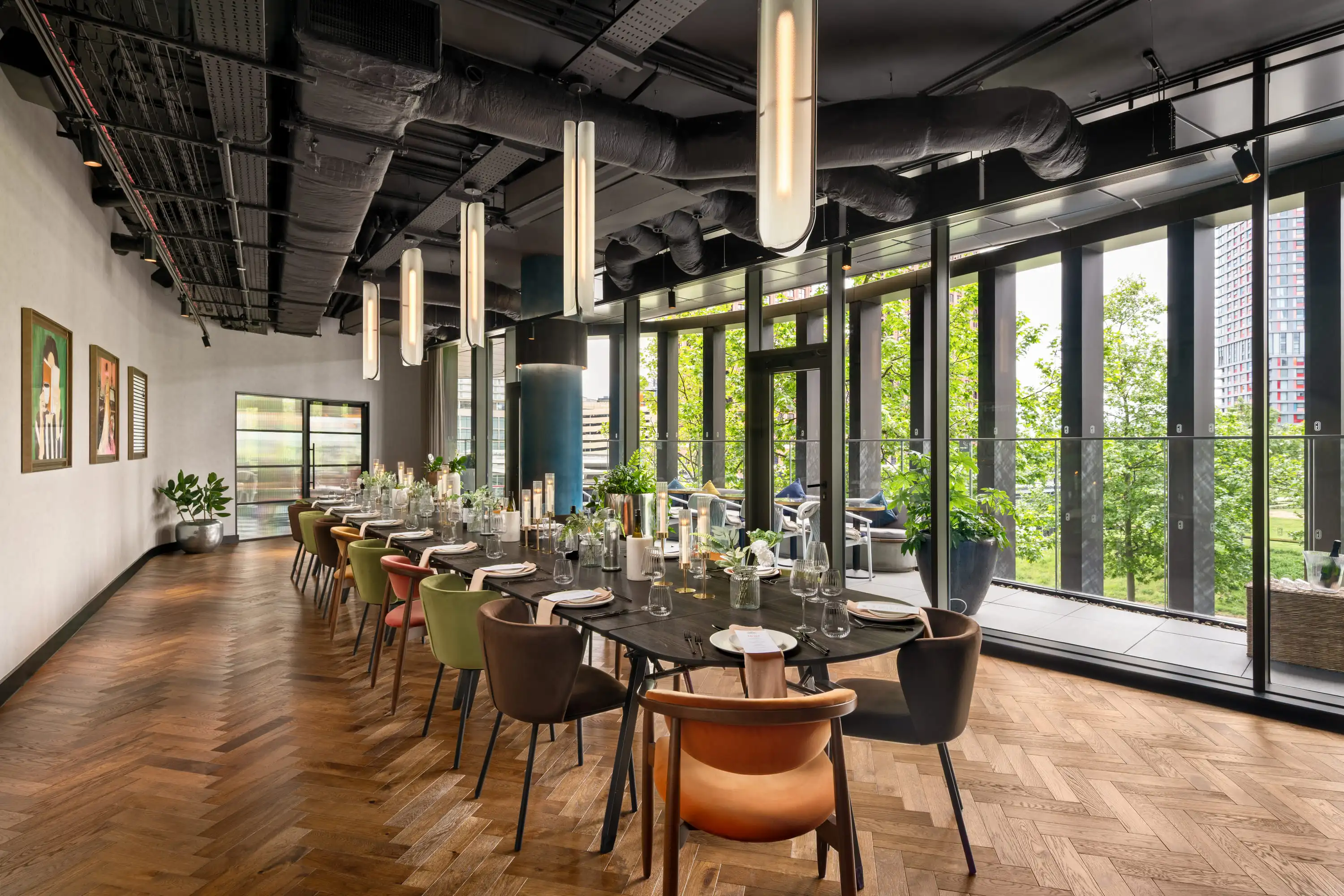 Modern dining room with long table, colorful chairs, hanging lights, and floor-to-ceiling windows overlooking greenery.