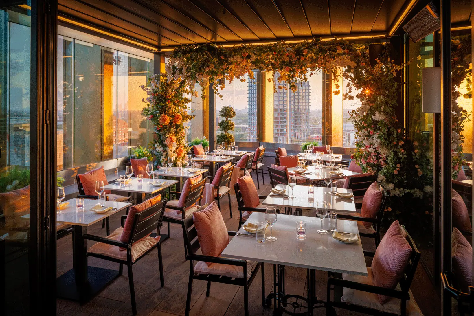 Cozy rooftop dining area at sunset with tables set with glassware and napkins, featuring chairs with pink cushions and floral decorations hanging from the ceiling and around columns, overlooking a cityscape with high-rise buildings through glass walls.