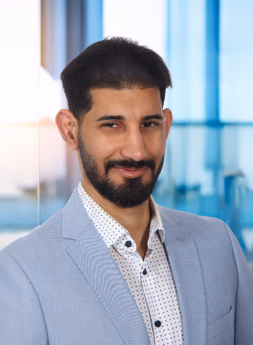 Portrait eines lächelnden Mannes mit dunklem Bart und mittellangem Haar, der einen hellblauen Anzug und ein weißes Hemd mit Muster trägt.