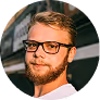 Portrait of a young man with glasses, short hair, and a beard, wearing a white shirt, with a street background