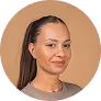 Headshot of a woman with straight dark hair pulled back, wearing small hoop earrings and a black top, against a beige background