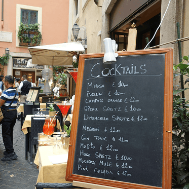 Tafel mit Cocktailmenü vor einem Straßencafé mit Gästen und Tischdekoration.